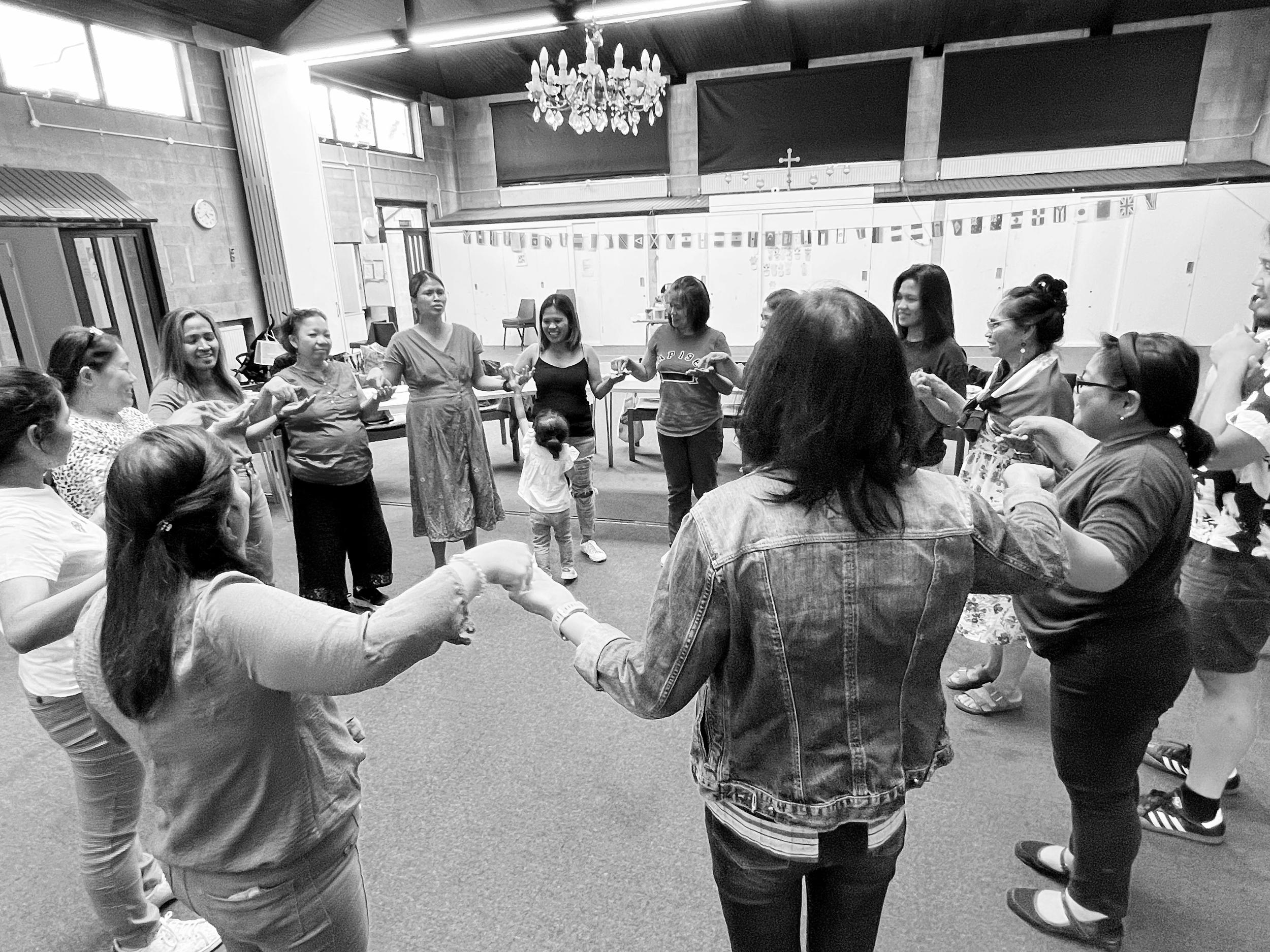 A group of diverse women and children holding hands in a circle inside a community room, engaging in a group activity.