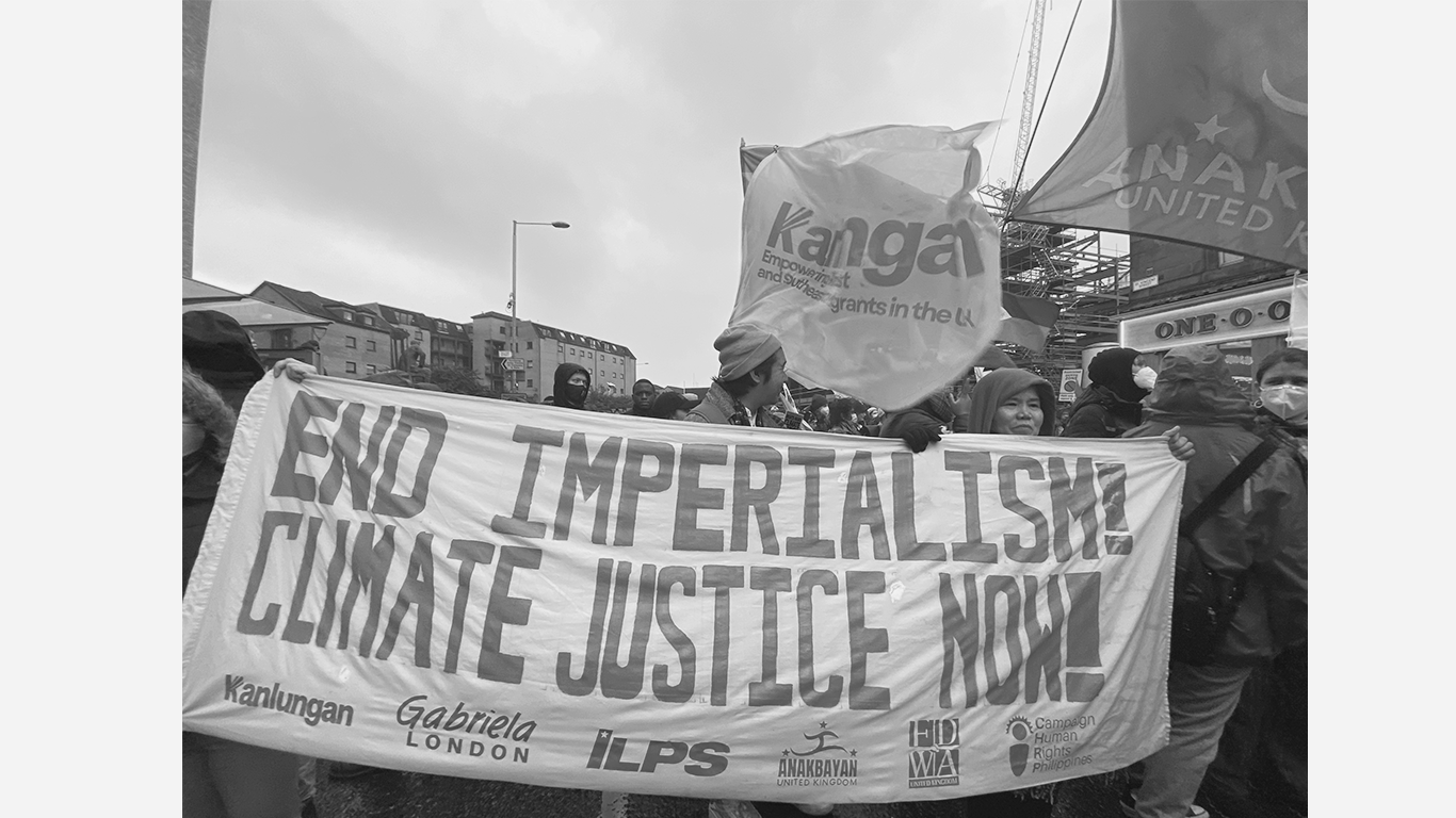 People participating in a climate protest holding a large banner that says, 'End Imperialism! Climate Justice Now!' Several individuals wear face masks, and some carry flags, including one with the word 'Kangana.' The protest takes place outdoors in an urban area with buildings and a cloudy sky in the background.