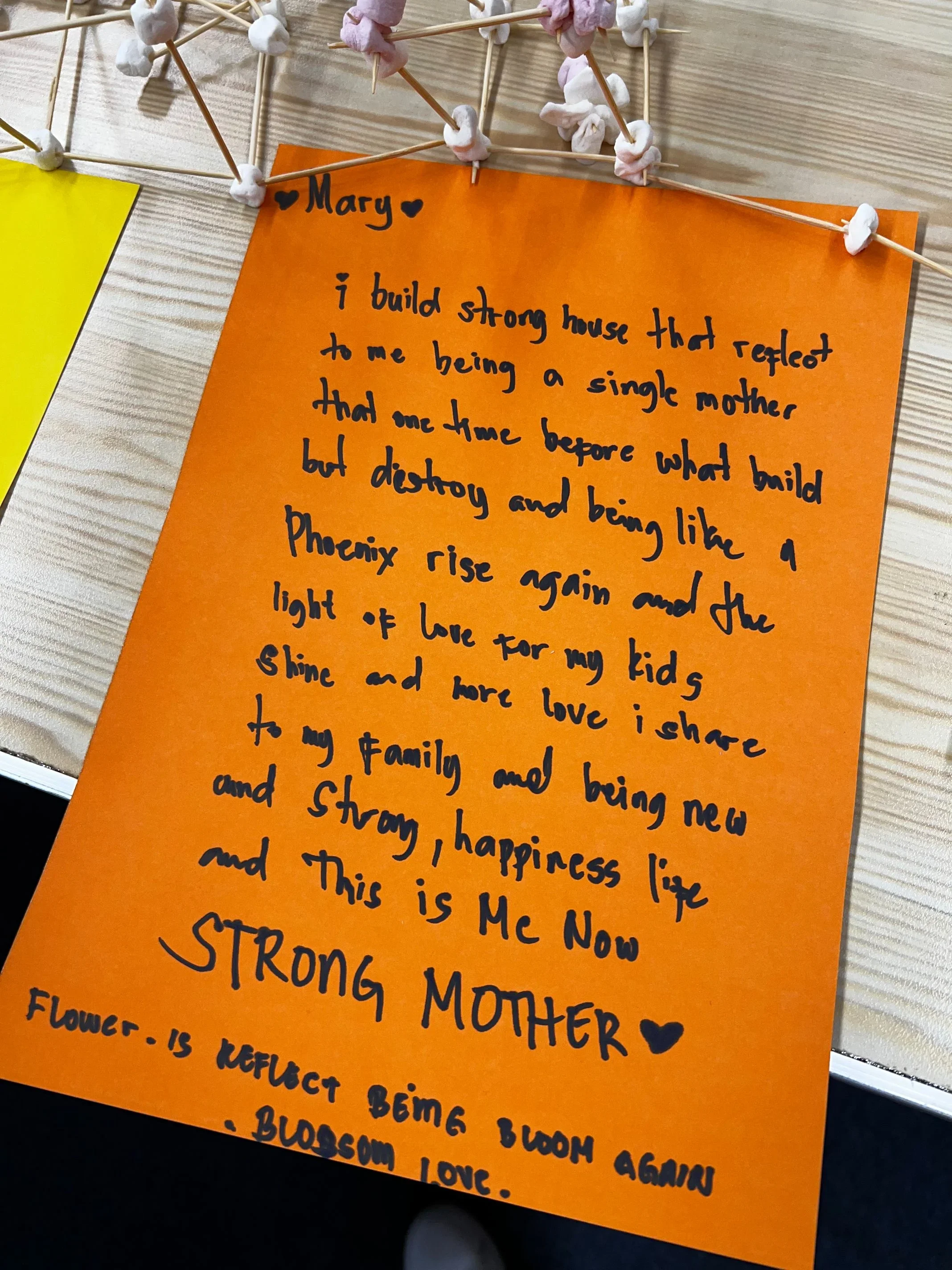Close-up of an orange handwritten note pasted on a wooden surface, with marshmallow skewers attached at the top, containing pink and white marshmallows.