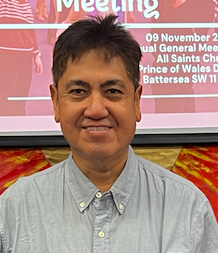 A man with short black hair, wearing a light gray button-up shirt, smiling at the camera in front of a pink event banner.