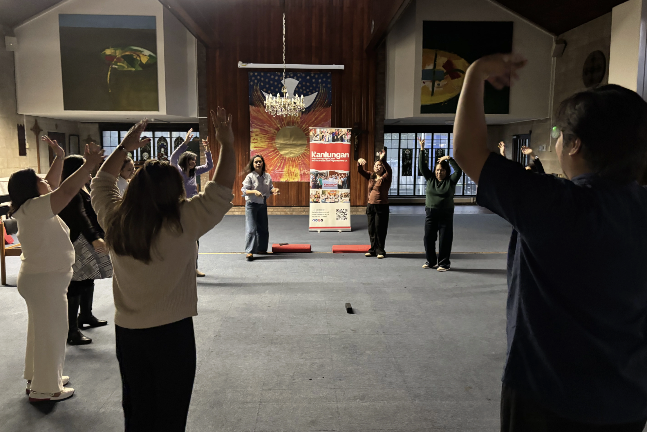 A group of people participating in a Tai Chi or similar exercise class inside a room with high windows, wooden paneling, a chandelier, and religious symbols on the walls. They are raising their arms and focusing on their movements.