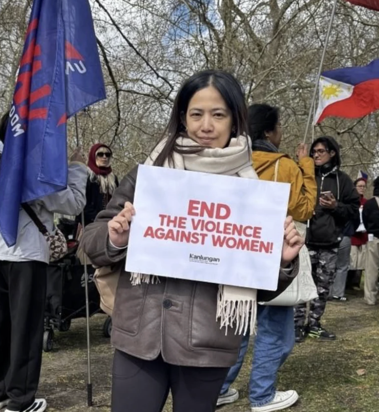 Woman holding a sign that reads 'End the violence against women!' at a protest or rally. Several other people are visible in the background, some holding flags.