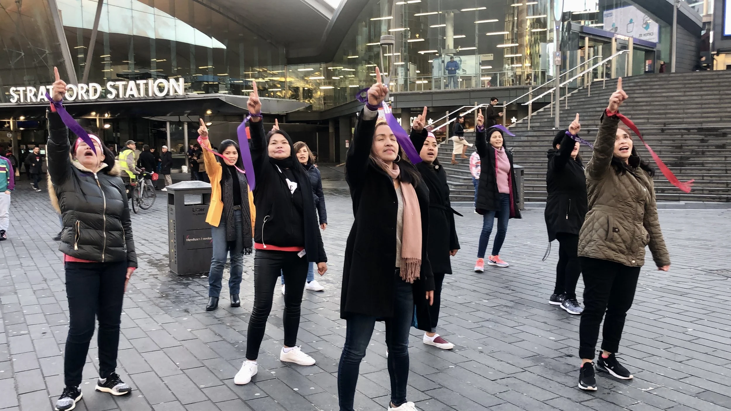 A group of diverse women standing on a city sidewalk, raising one finger in a gesture of unity or protest, in front of the Stratford Station.