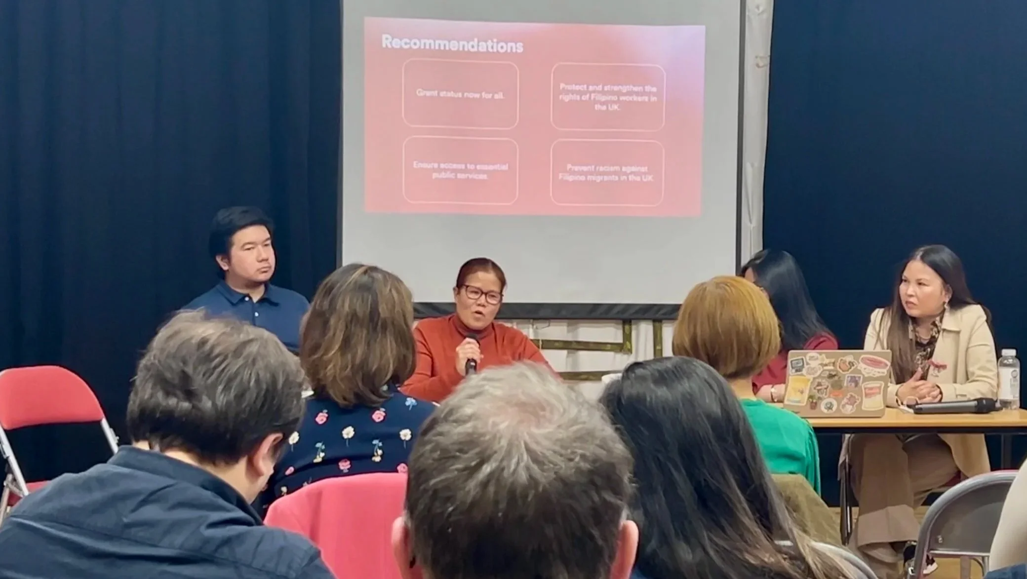 A panel of four people seated at a table in front of an audience during a presentation or discussion. A woman in the center is speaking into a microphone. Behind them, a projection screen displays a slide with the heading 'Recommendations' and four bullet points. The audience members are seen from behind, with some taking notes or listening attentively.