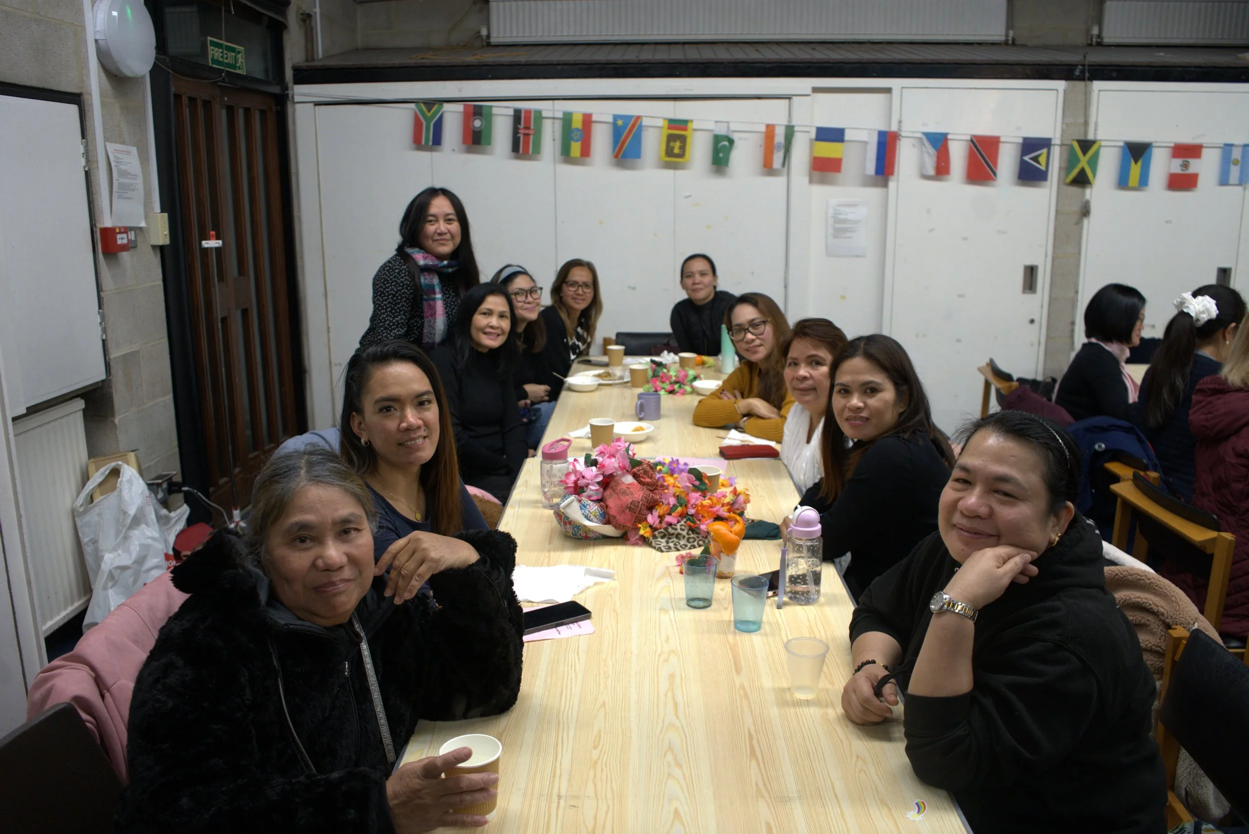 Group of women sitting around a long table decorated with flowers, in a room with international flags hanging on the wall.