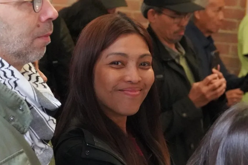 A group of people standing close together, with a woman smiling at the camera in the center. The woman has long dark hair and is wearing a black jacket. The background includes brick walls and other individuals, some looking at their phones.