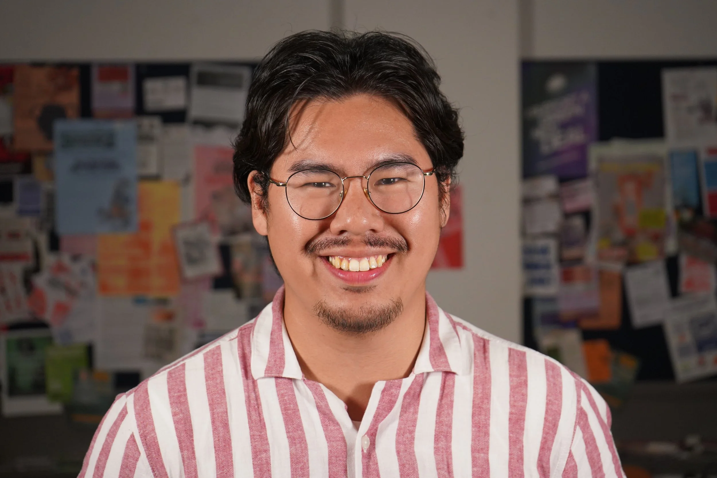 A young man with black hair, glasses, and a goatee smiling, wearing a pink and white striped shirt, standing in front of a bulletin board filled with various colored flyers and notices.