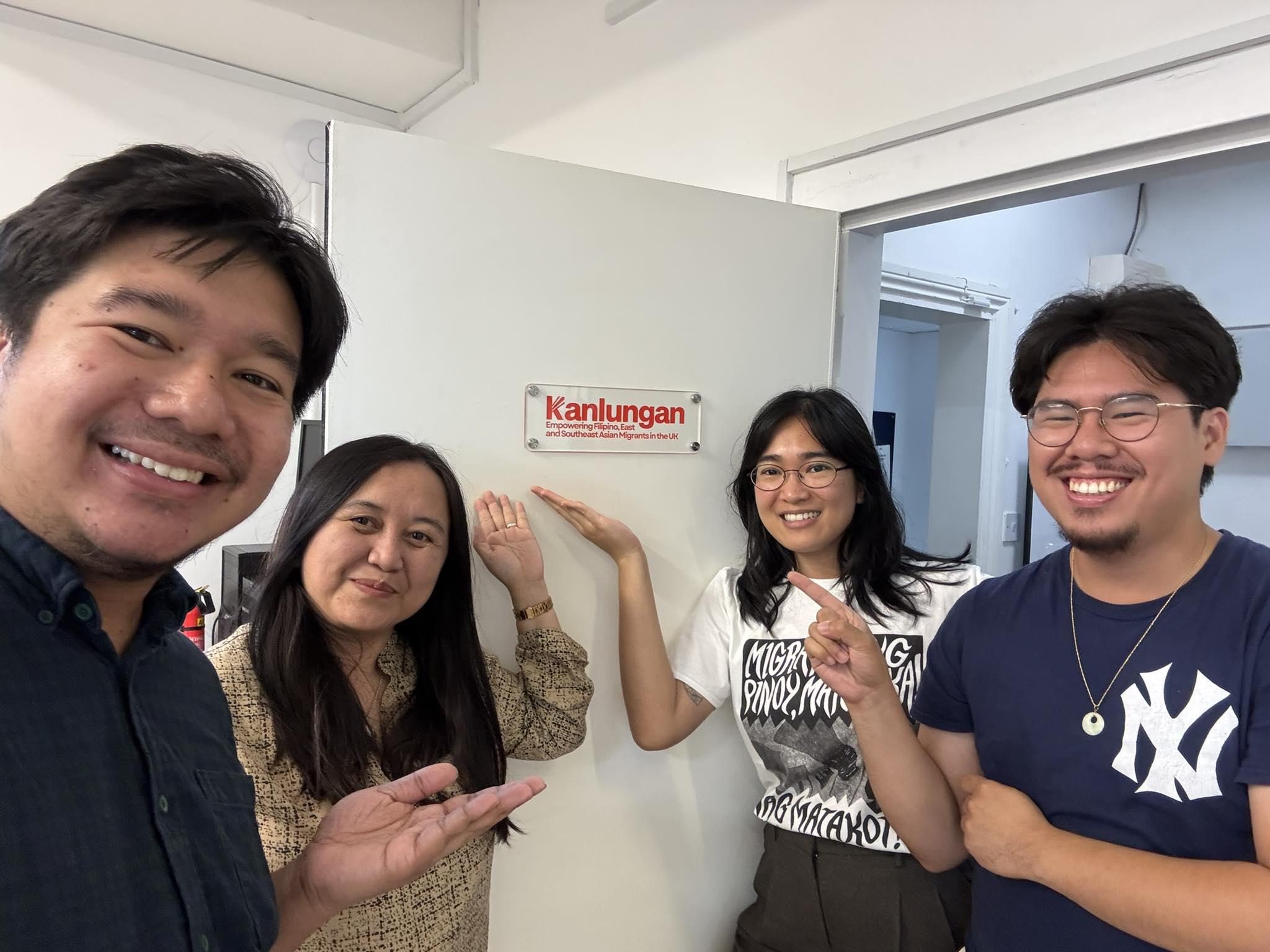 Group of five people smiling and pointing at a sign on the wall that reads 'Kanlungan Empowering Filipino, East and Southeast Asian Migrants in the UK.'