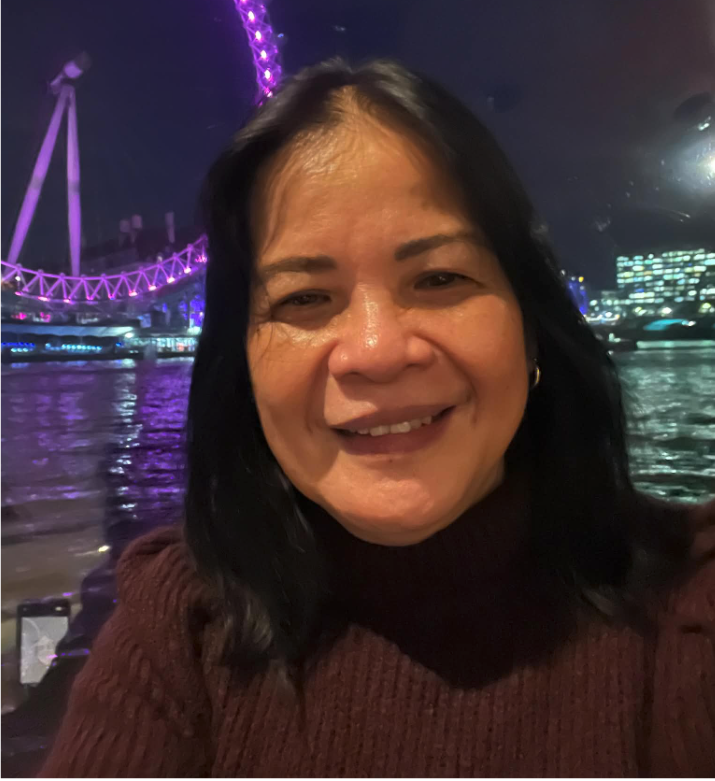 A woman smiling at night with a lit Ferris wheel and cityscape in the background, next to a body of water.