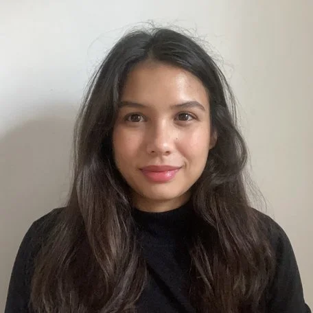 A young woman with long dark hair and a black shirt smiling against a plain white wall.