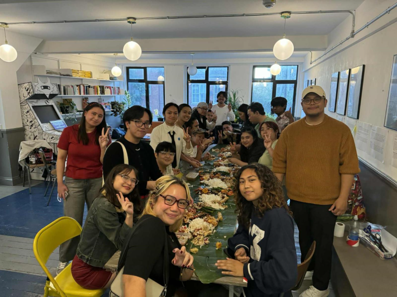 Group of diverse people gathered around a long table with food, in a well-lit room with windows, bookshelves, and artwork.