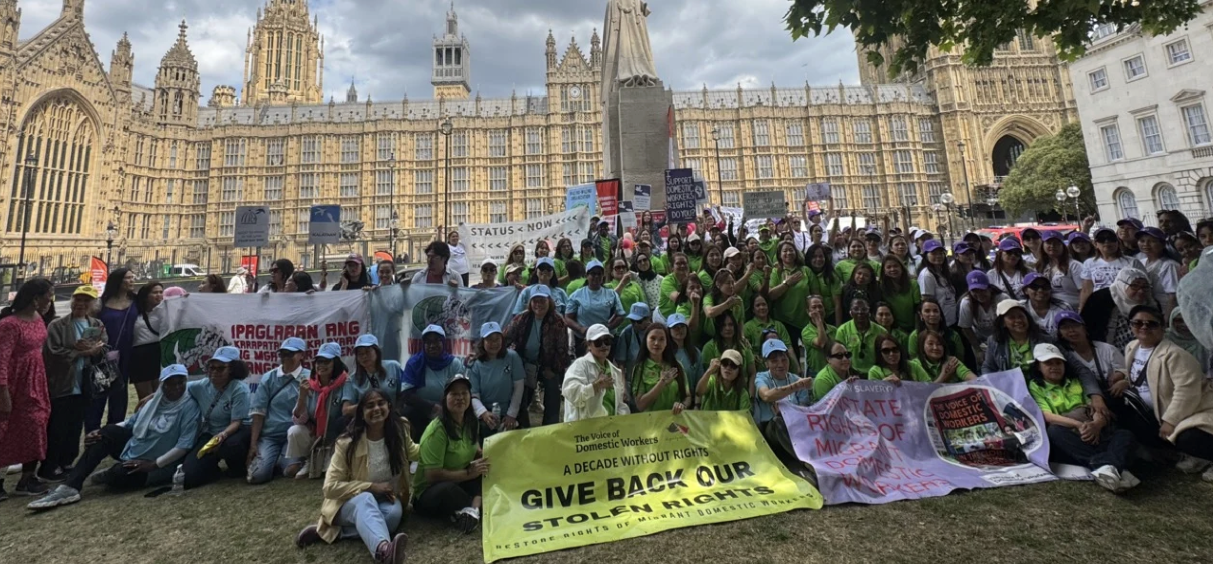 A large group of people gathered in front of a historic building, holding signs and banners advocating for the rights of domestic workers. The crowd includes men and women wearing various colorful clothes and hats, and some are smiling at the camera.