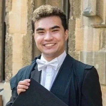 Young man in a graduation gown and cap smiling outside a stone building.