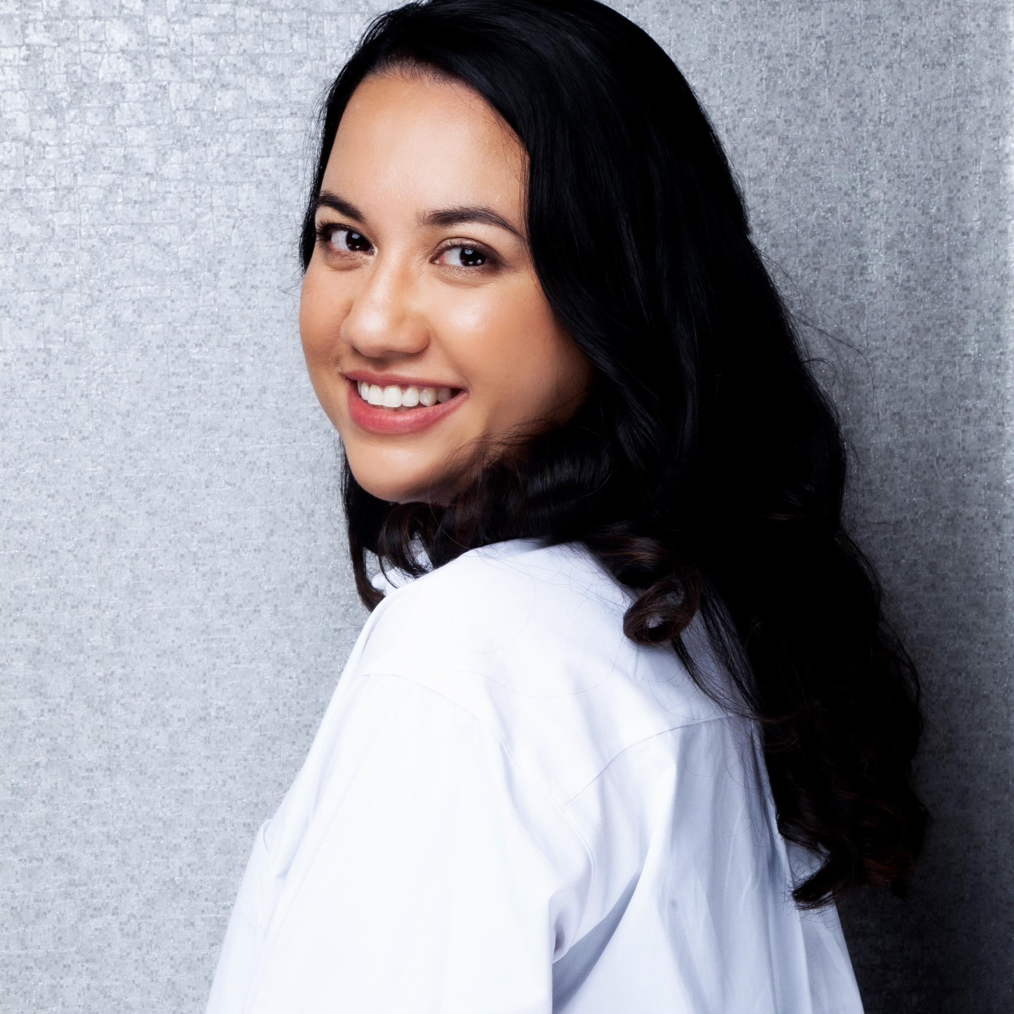 Close-up of a smiling woman with long dark hair, wearing a white shirt, standing against a light gray textured background.