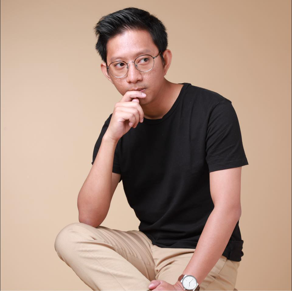 A young man with black hair and glasses, wearing a black T-shirt and beige pants, sitting with his knee up, touching his chin thoughtfully against a beige background.