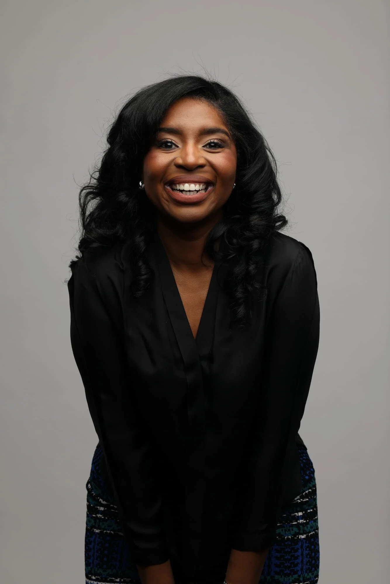 Rashida is smiling, wearing a black blouse and patterned skirt, against a plain gray background.