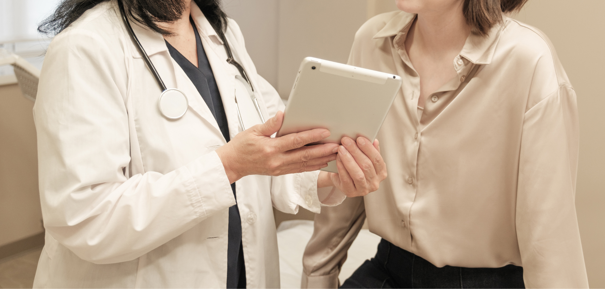 A healthcare professional, wearing a white coat and stethoscope, shows a tablet to a patient in a beige shirt in a clinic.