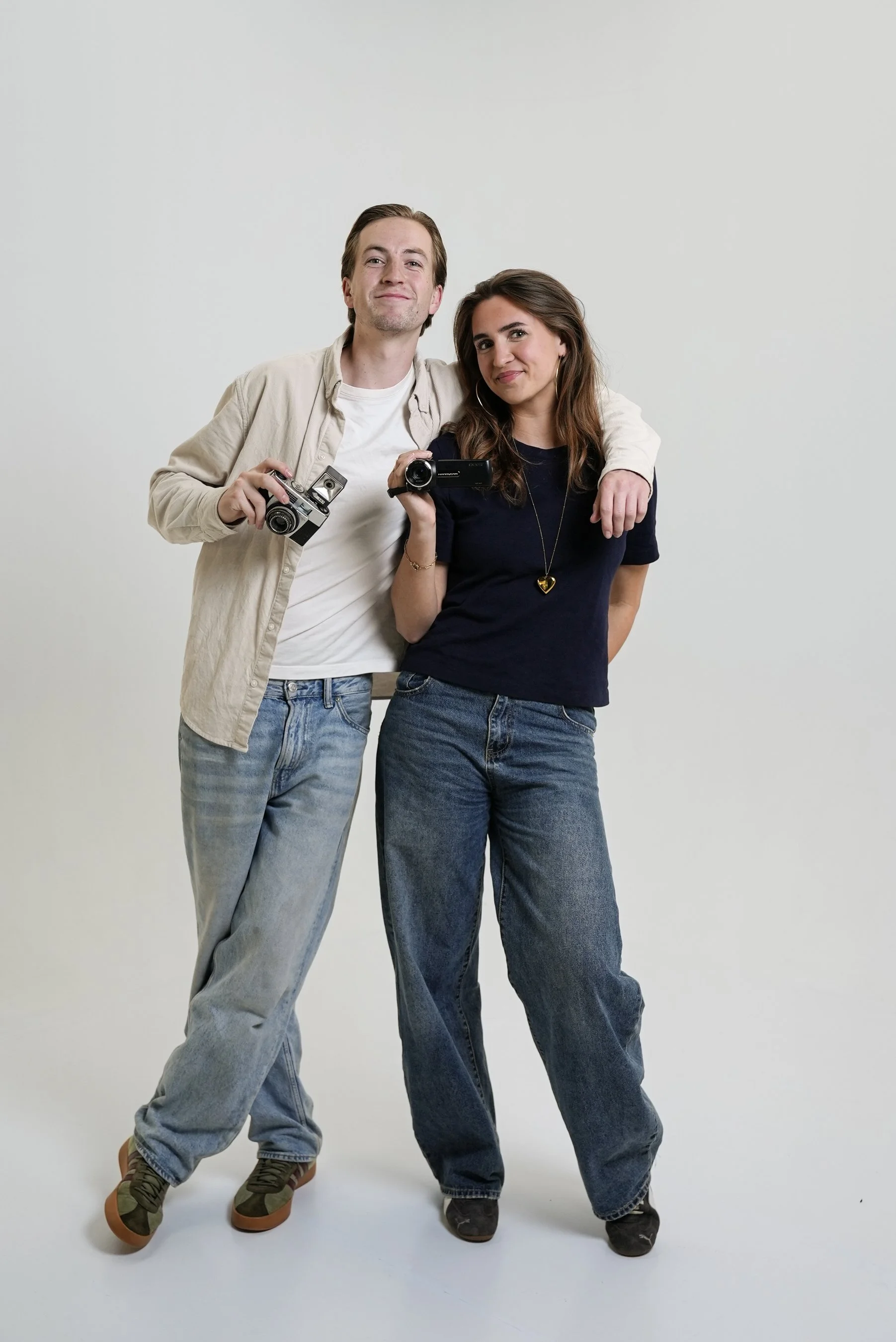 Een man en een vrouw staan naast elkaar, de man heeft zijn arm om de schouders van de vrouw. Ze lachen en houden camera's in de handen. Ze dragen casual kleding en staan voor een effen witte achtergrond.