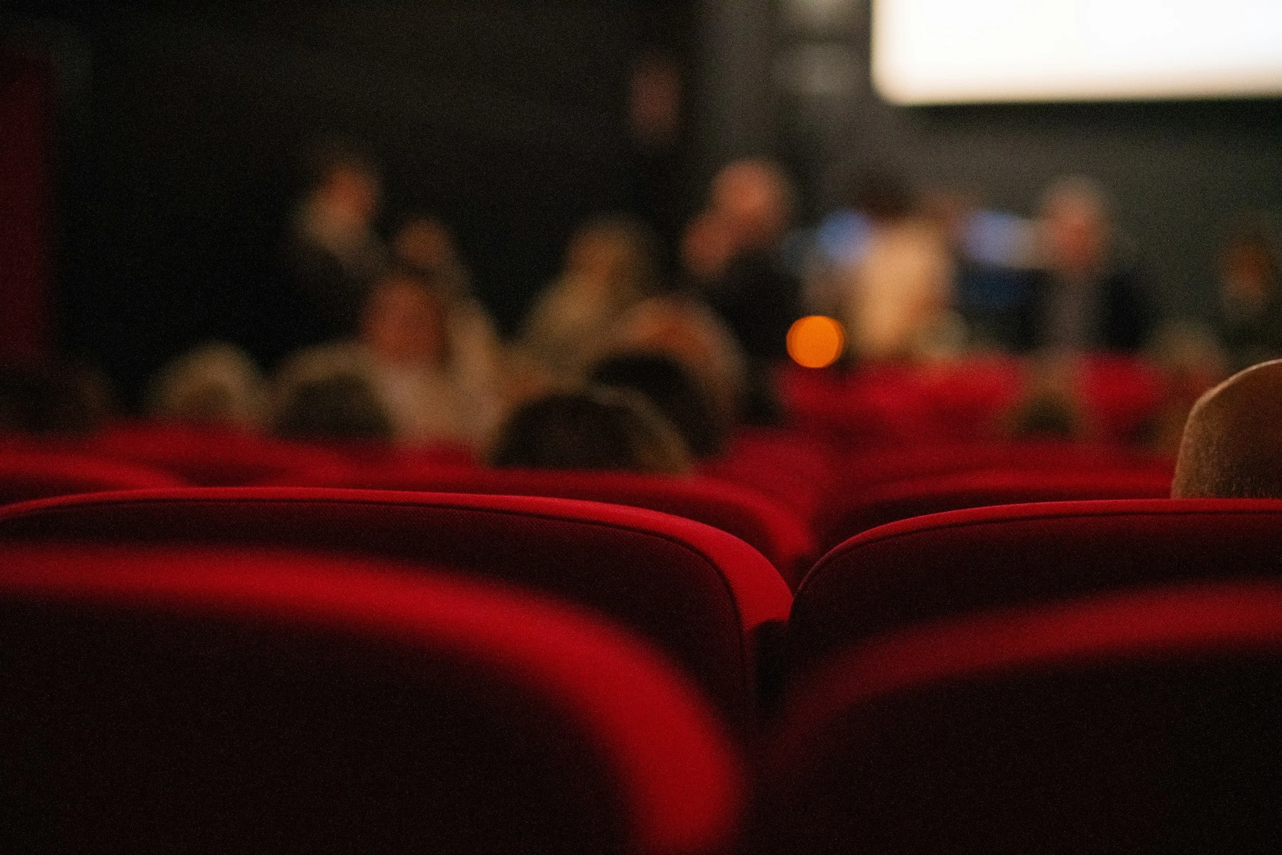Blurred image of theater seats with a dark background, illuminated by faint lights, and a large white screen or light in the upper right corner.