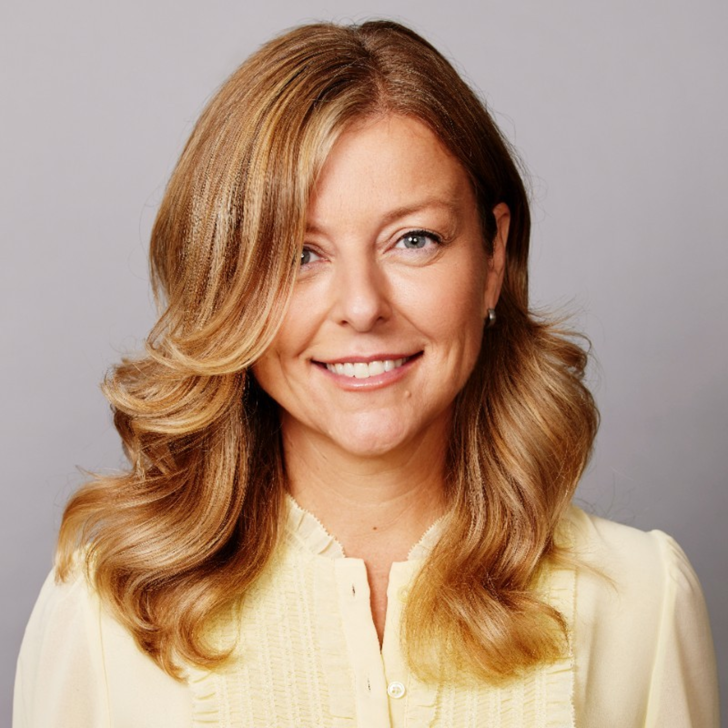 A woman with long, wavy blonde hair, blue eyes, and a bright smile, wearing a light-colored blouse, against a plain gray background.