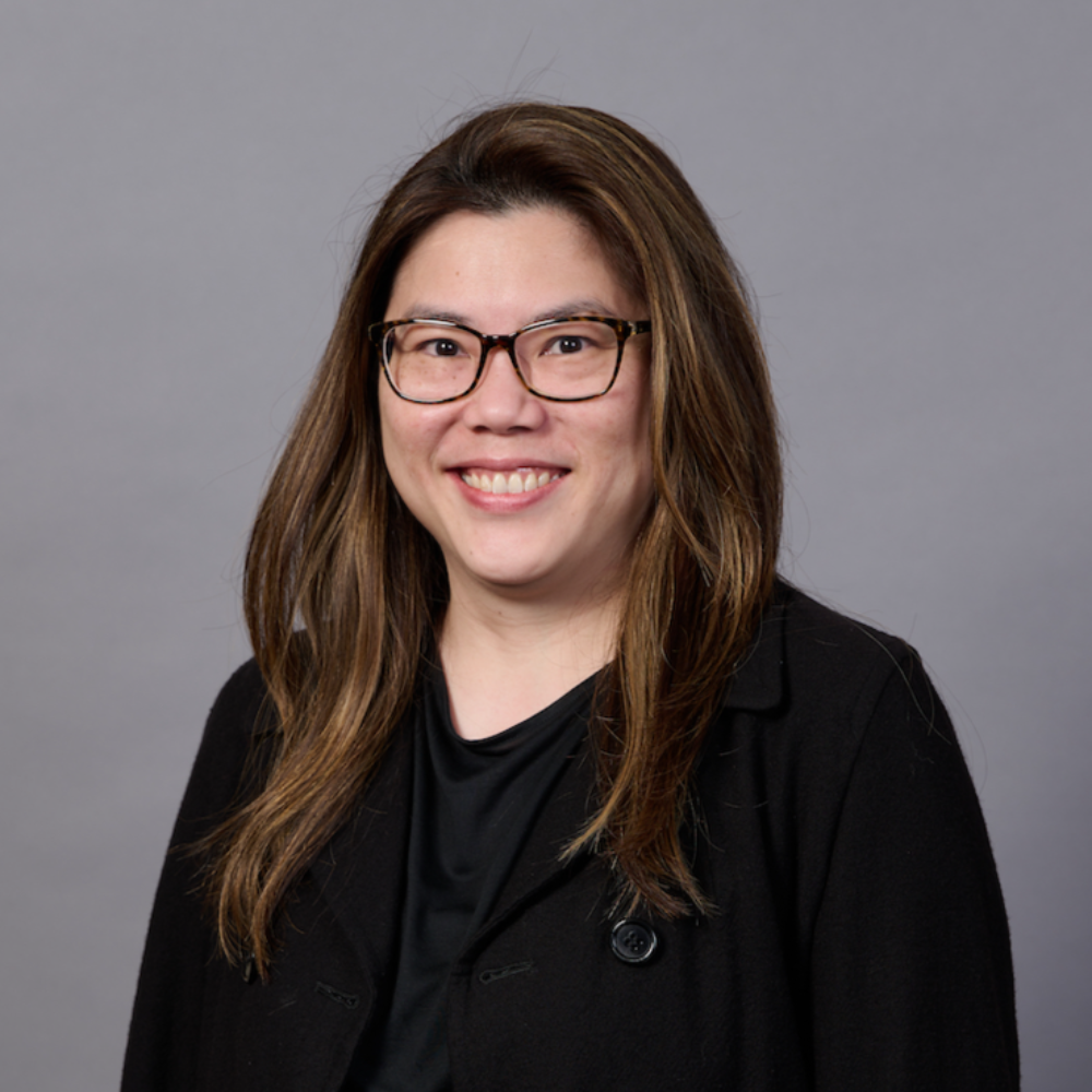 A woman with long brown hair, glasses, and a black blazer smiling in front of a gray background.