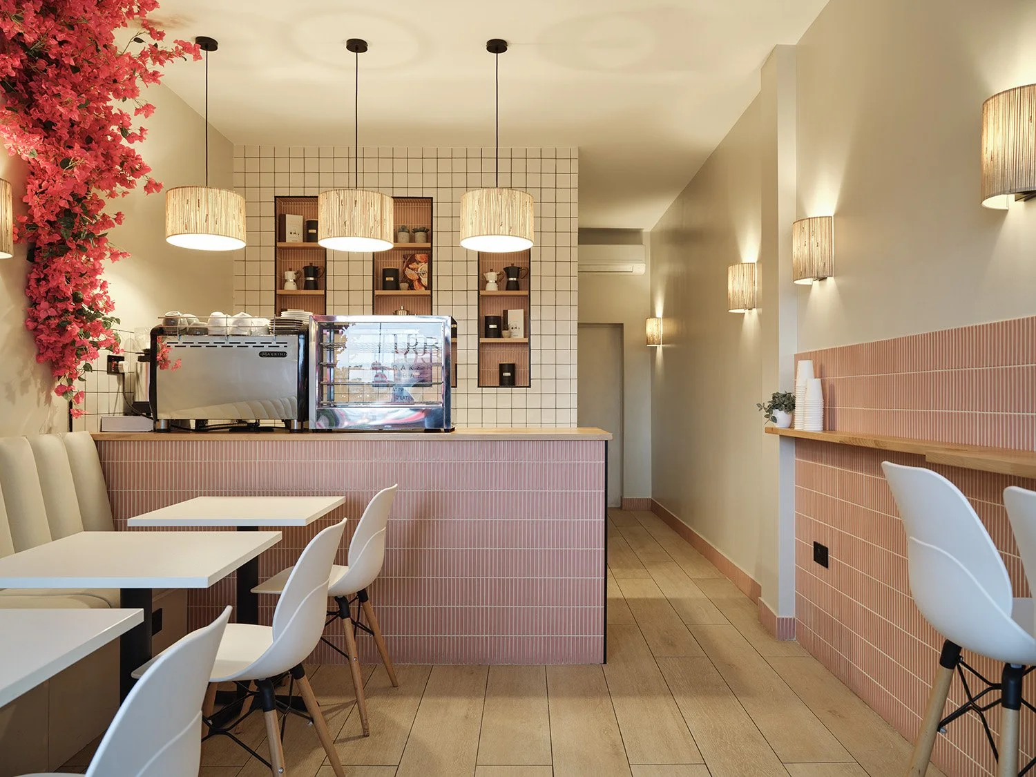 Interior of a modern cafe with pink tiled counter, white chairs, wall-mounted lamps, decorative shelves, and a coffee machine, with pink flowers on the wall.
