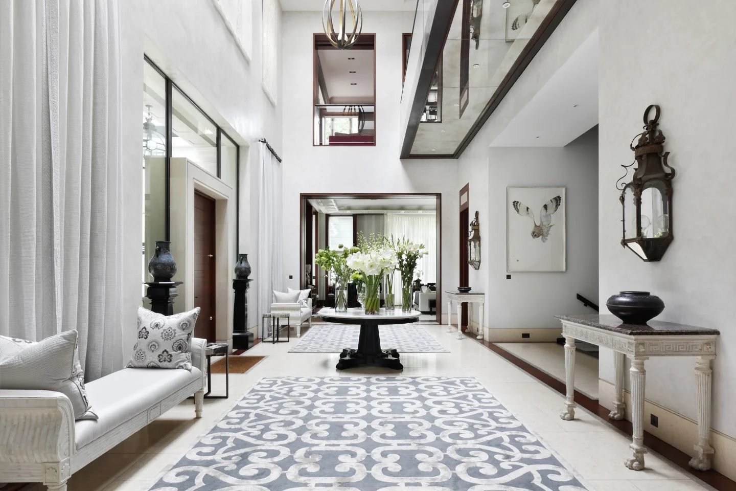 Spacious, elegant living room with high ceilings, white walls, large windows with white curtains, a central round table with flower vases, a white sofa with decorative pillows, a patterned area rug, and wall-mounted vintage lanterns.