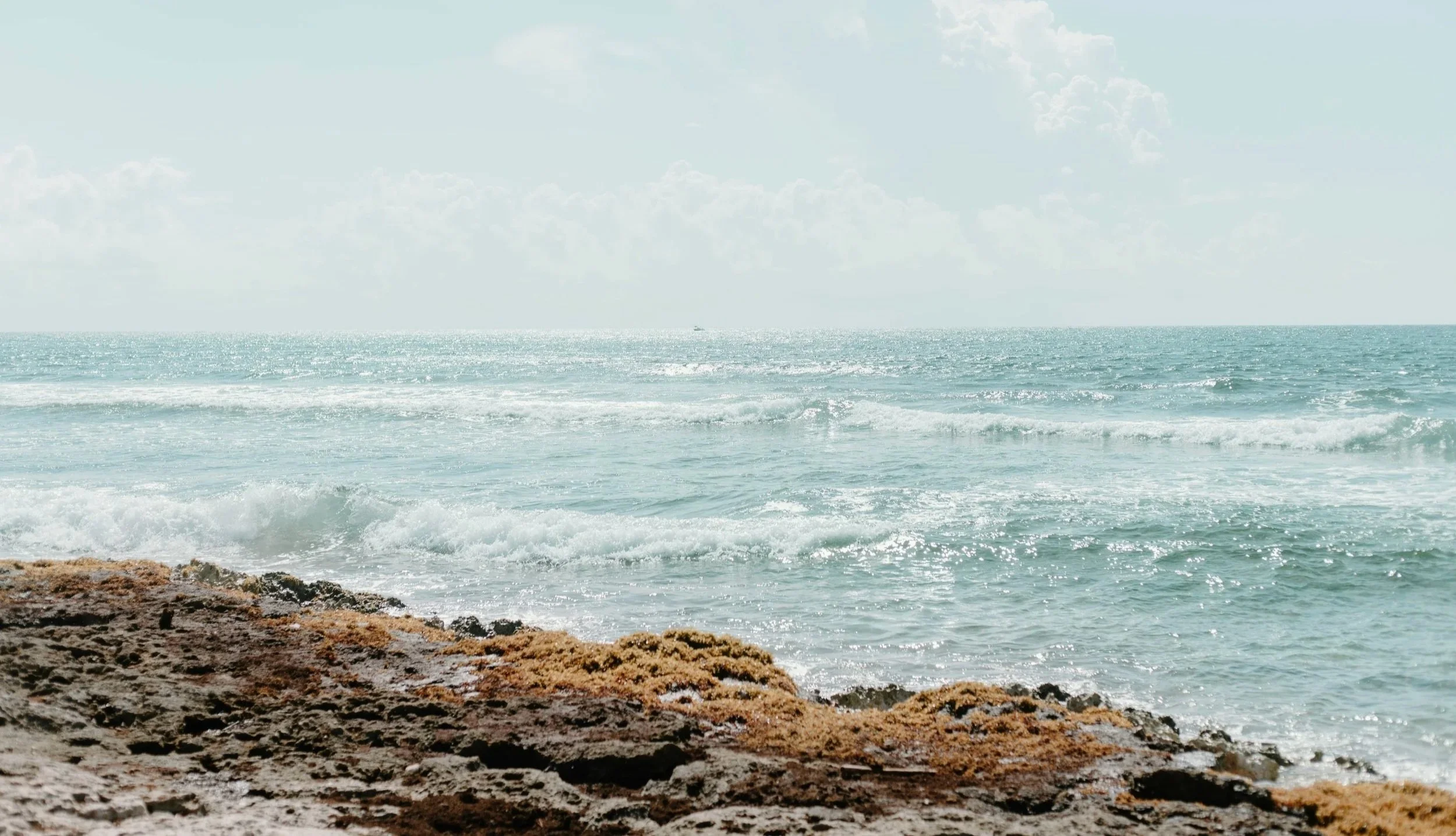 View of the ocean with waves crashing on a sandy and rocky shoreline under a cloudy sky.