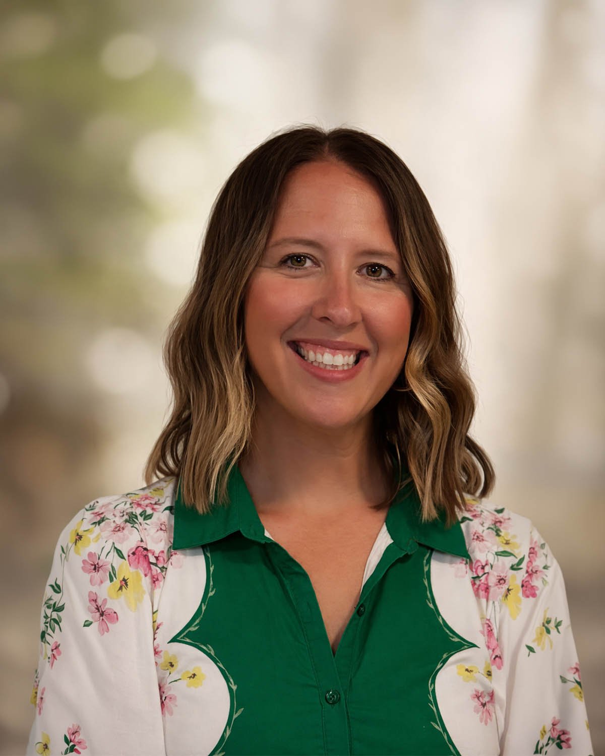 A woman with shoulder-length wavy brown hair smiling, wearing a green and white floral shirt, with a blurred background.