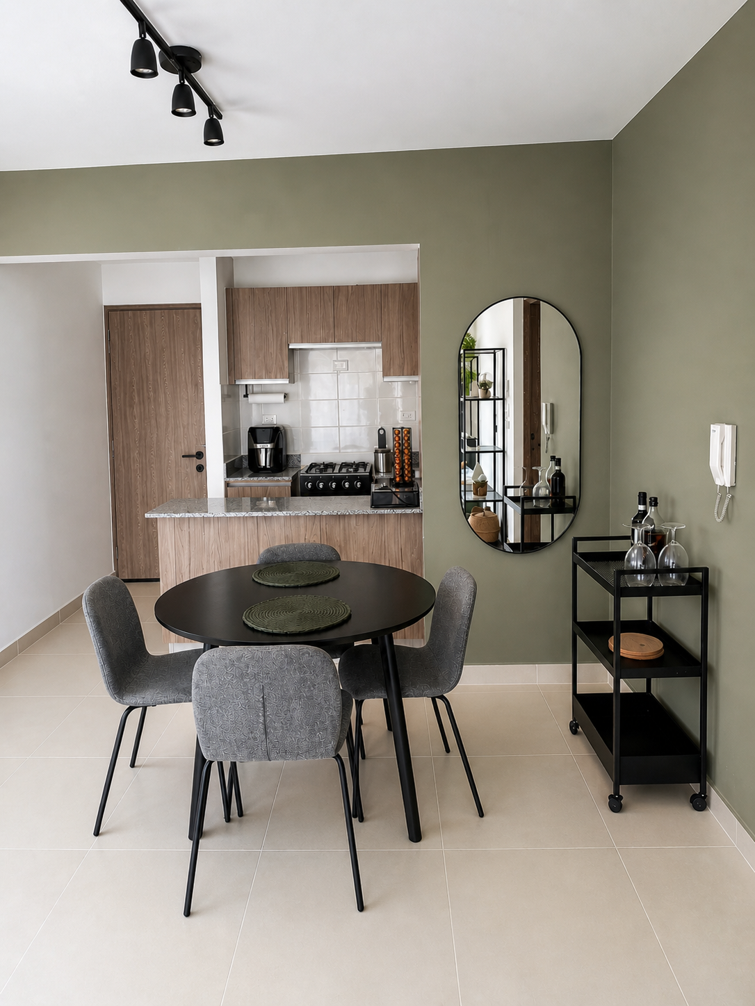 Interior view of a modern dining room and kitchen with a black round table, four gray upholstered chairs, a black rolling bar cart with wine bottles and glasses, a tall mirror, and wooden kitchen cabinets, walls are painted green and off-white, and t