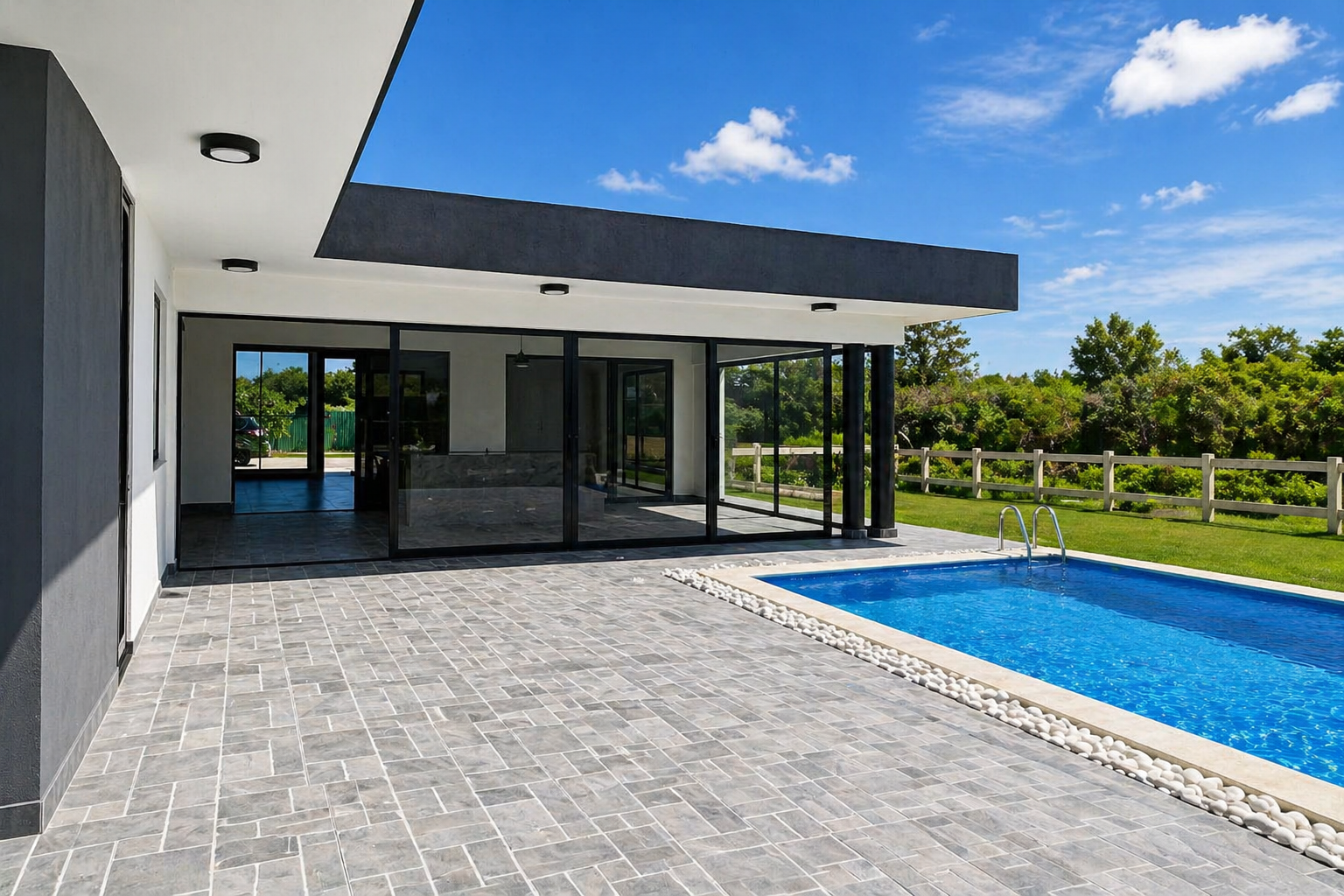 Modern house with large glass sliding doors, outdoor patio, swimming pool with white stones border, green lawn, wooden fence, trees, and partly cloudy blue sky.