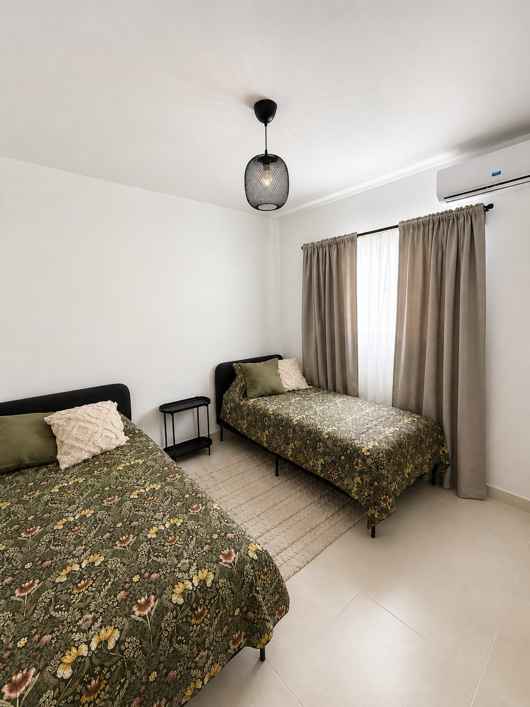 Two single beds with floral-patterned bedding and pillows, a window with beige curtains, a small black bedside table, a beige rug, white walls, tiled floor, ceiling light, and a wall-mounted air conditioner.