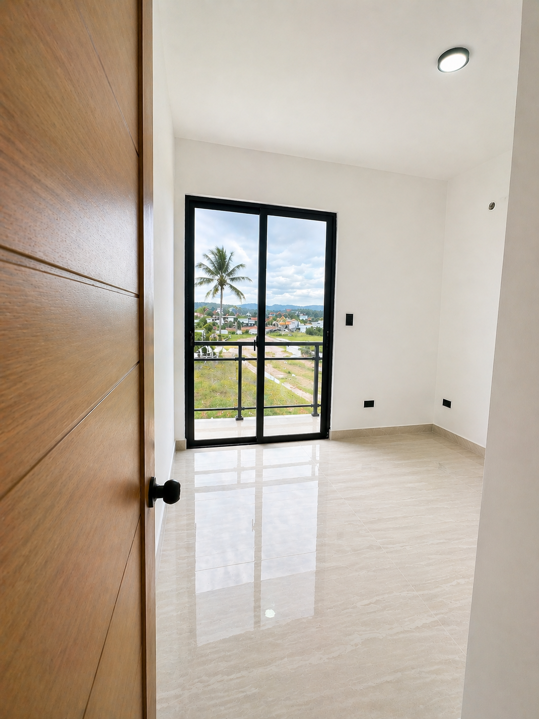 Empty room with a sliding glass door leading to a balcony, views of a tropical landscape with a palm tree and houses, wooden door partially open, glossy tiled floor, white walls, ceiling light.