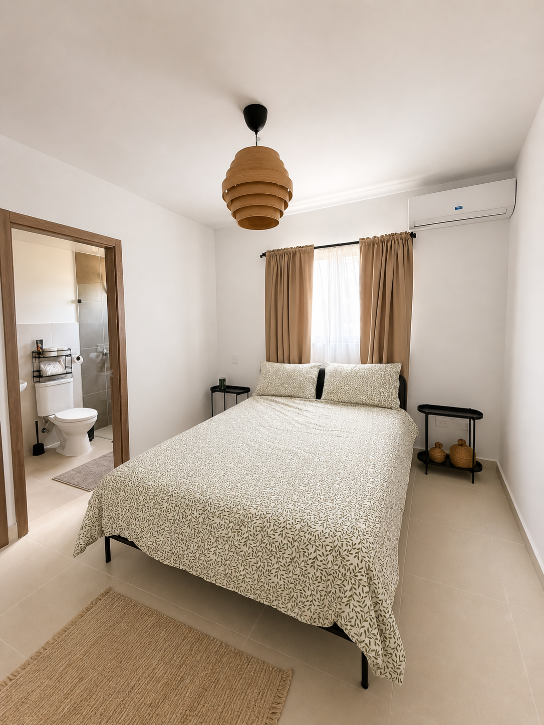 A cozy bedroom with a bed, beige curtains, a window, nightstands, and a view into a bathroom with a toilet and sink.
