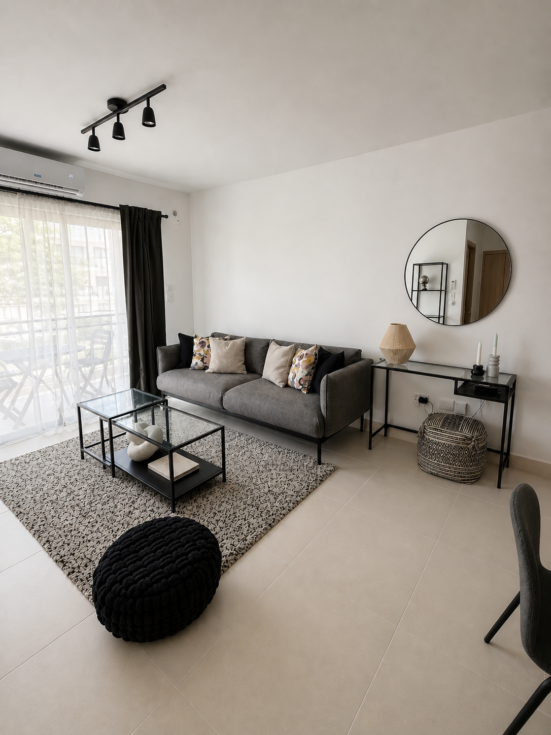 Modern living room with gray sofa, decorative pillows, glass-top coffee table, black knitted pouf, large window with sheer and black curtains, and minimalist decor including a round mirror, lamp, candles, and shelf.
