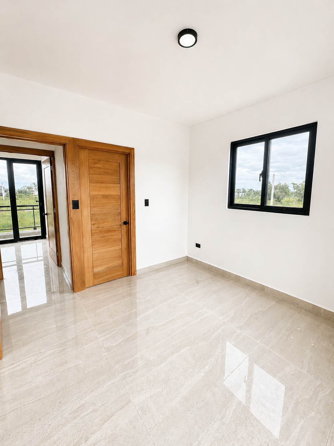 Empty room with white walls, beige tiled floor, a black-framed window showing greenery outside, a ceiling light, and a wooden door leading to a balcony or another room.