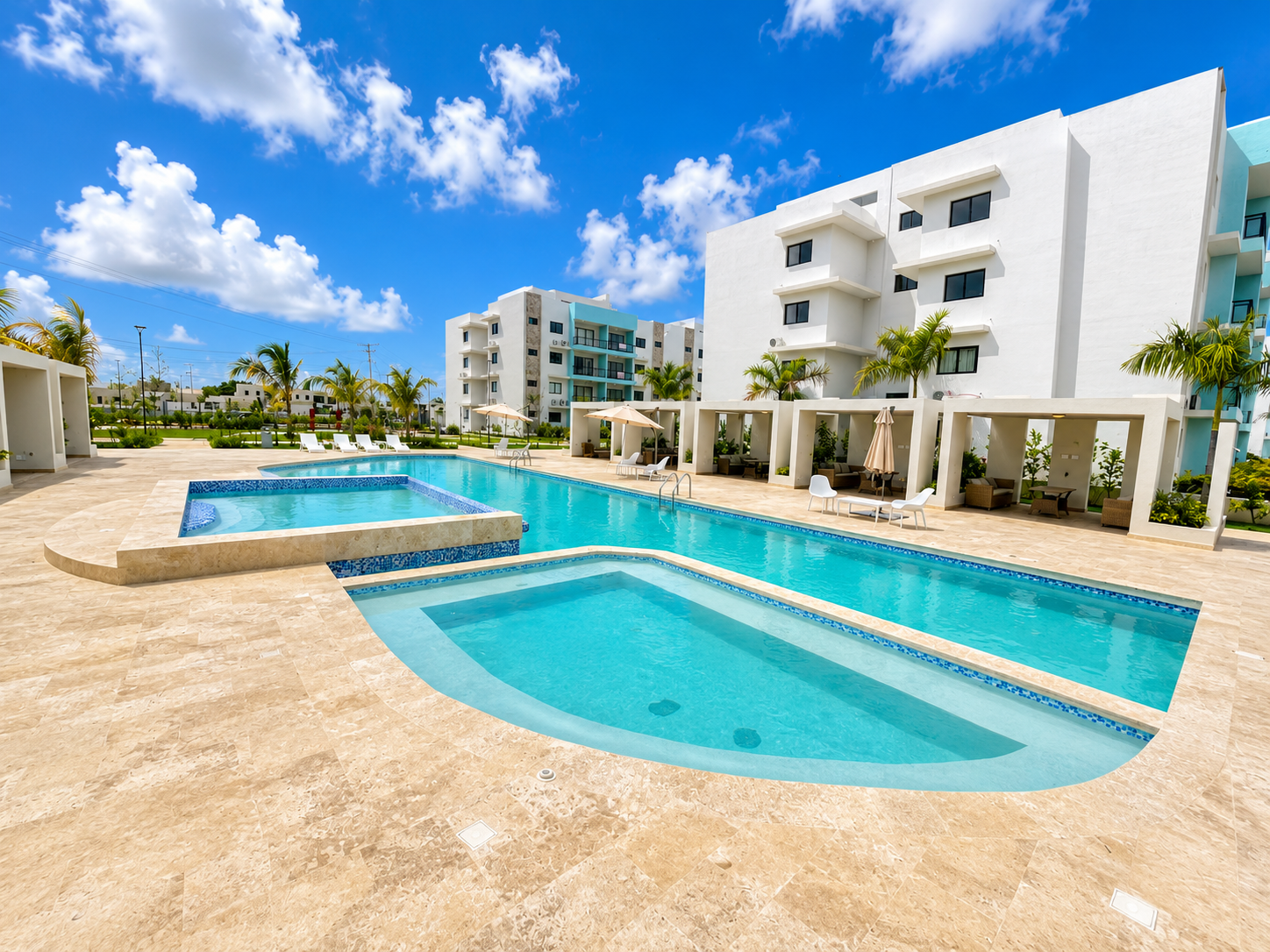 Luxury outdoor swimming pool area with lounge chairs, umbrellas, and modern white buildings in the background under a bright blue sky.