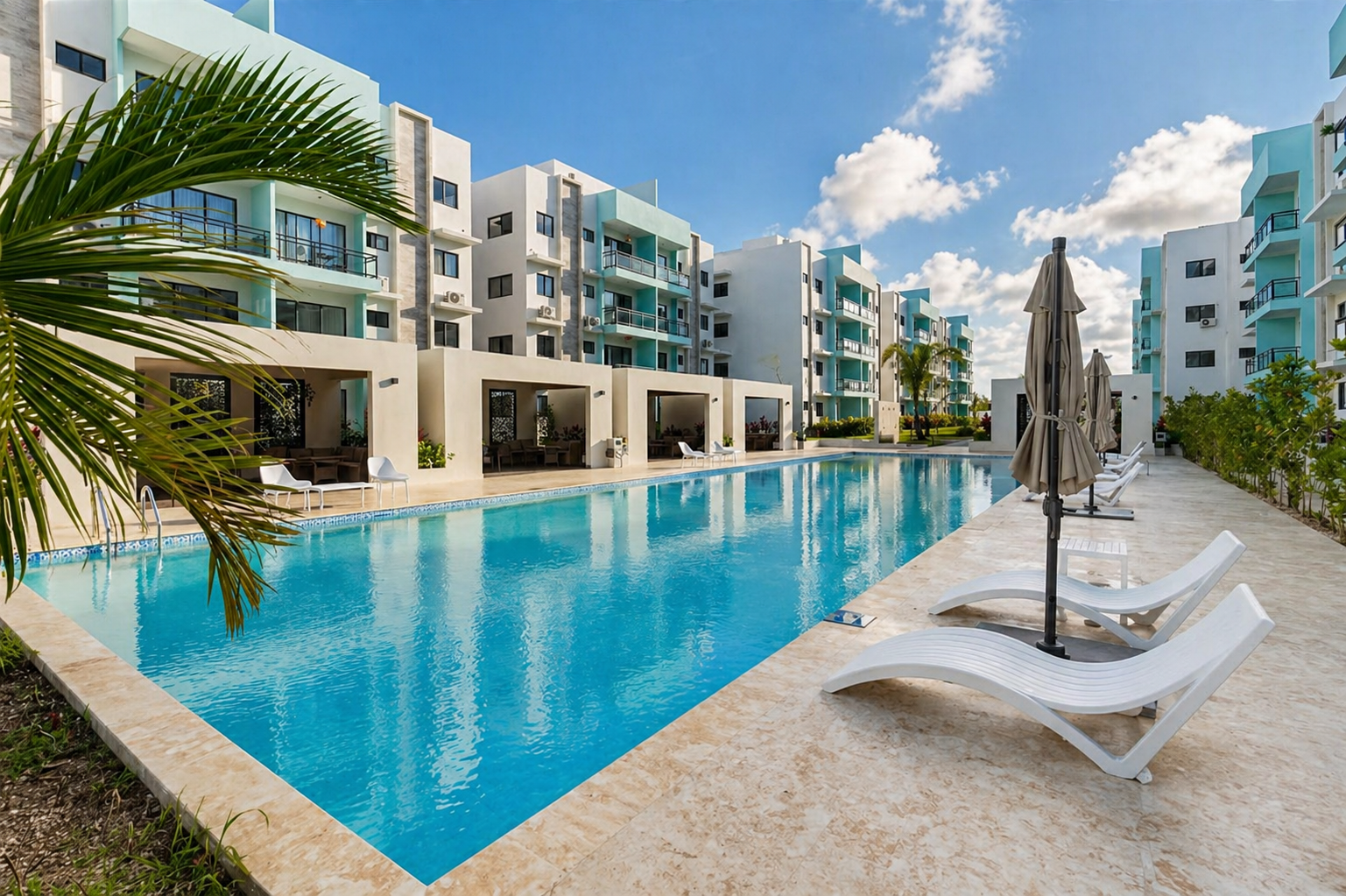 A modern hotel courtyard with a swimming pool, white lounge chairs, and umbrellas, surrounded by multi-story apartment buildings under a partly cloudy sky.