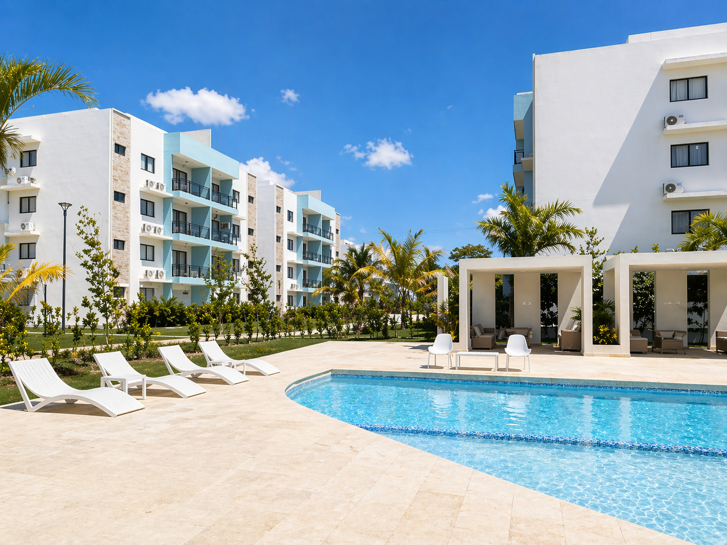 A resort with a swimming pool surrounded by lounge chairs, cabanas, and modern white apartment buildings under a blue sky with some clouds, with palm trees and greenery.