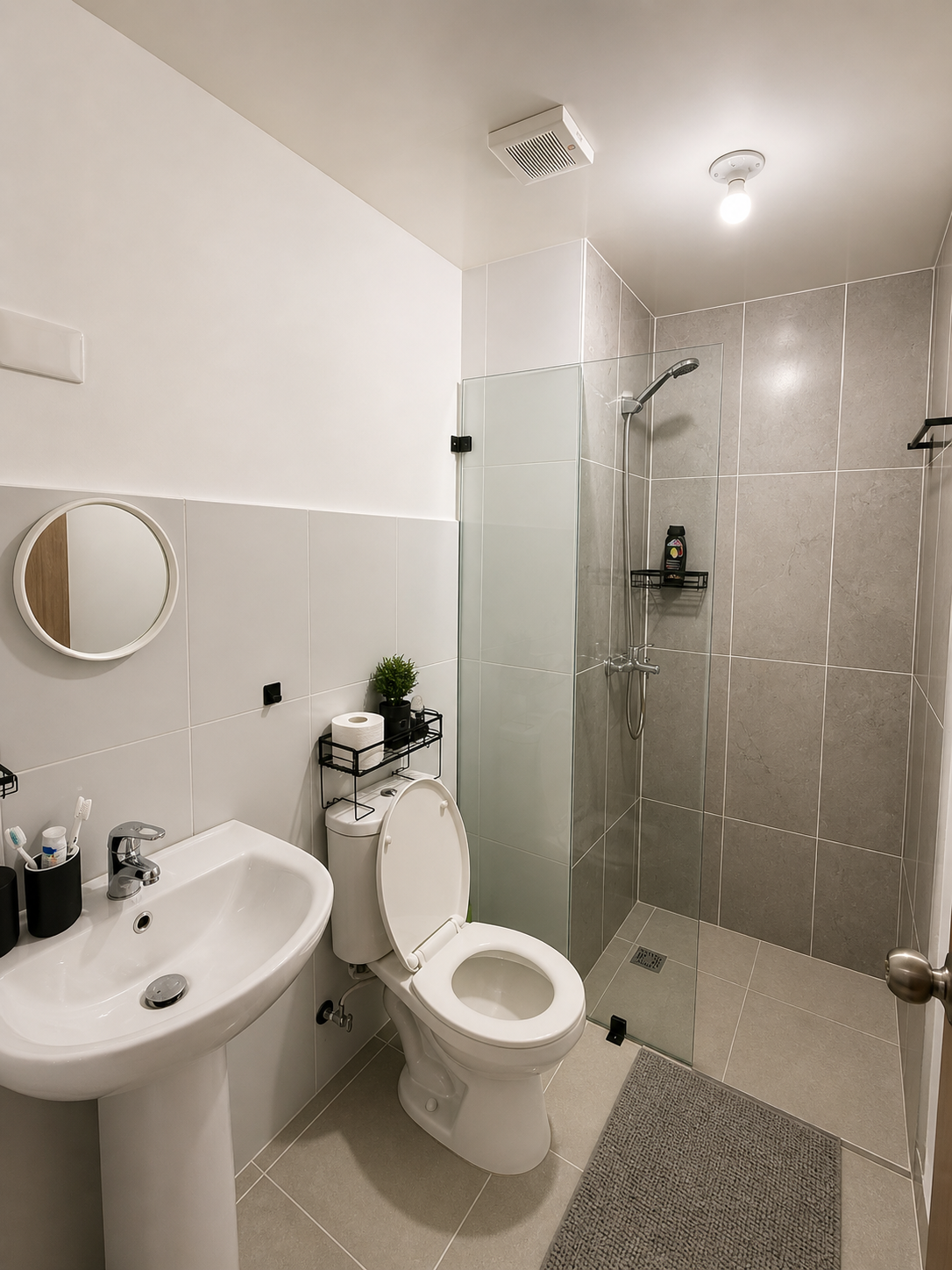 A modern, minimalist bathroom with a white pedestal sink, a round mirror, a toilet with a black metal shelf holding a plant and toilet paper, a clear glass shower with a black shelf and shower head, and a small floor mat. The walls are tiled half-hei
