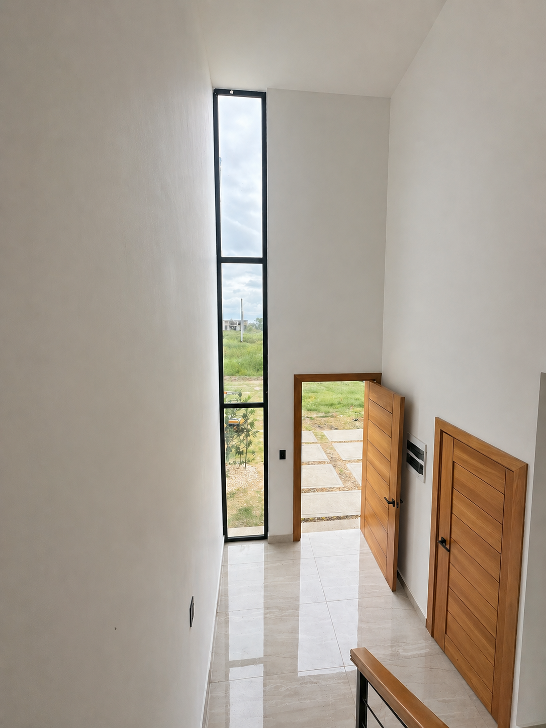 Interior view of a modern entrance with wooden doors, a tall vertical window, and a view of the outdoors with greenery and paved walkway.