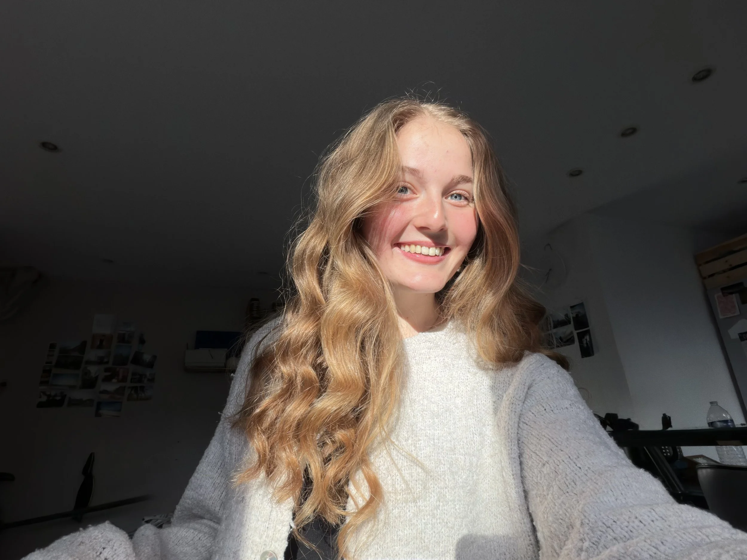 A young woman with long wavy blonde hair and light skin is smiling, looking at the camera in a room with wall art and a dark ceiling.