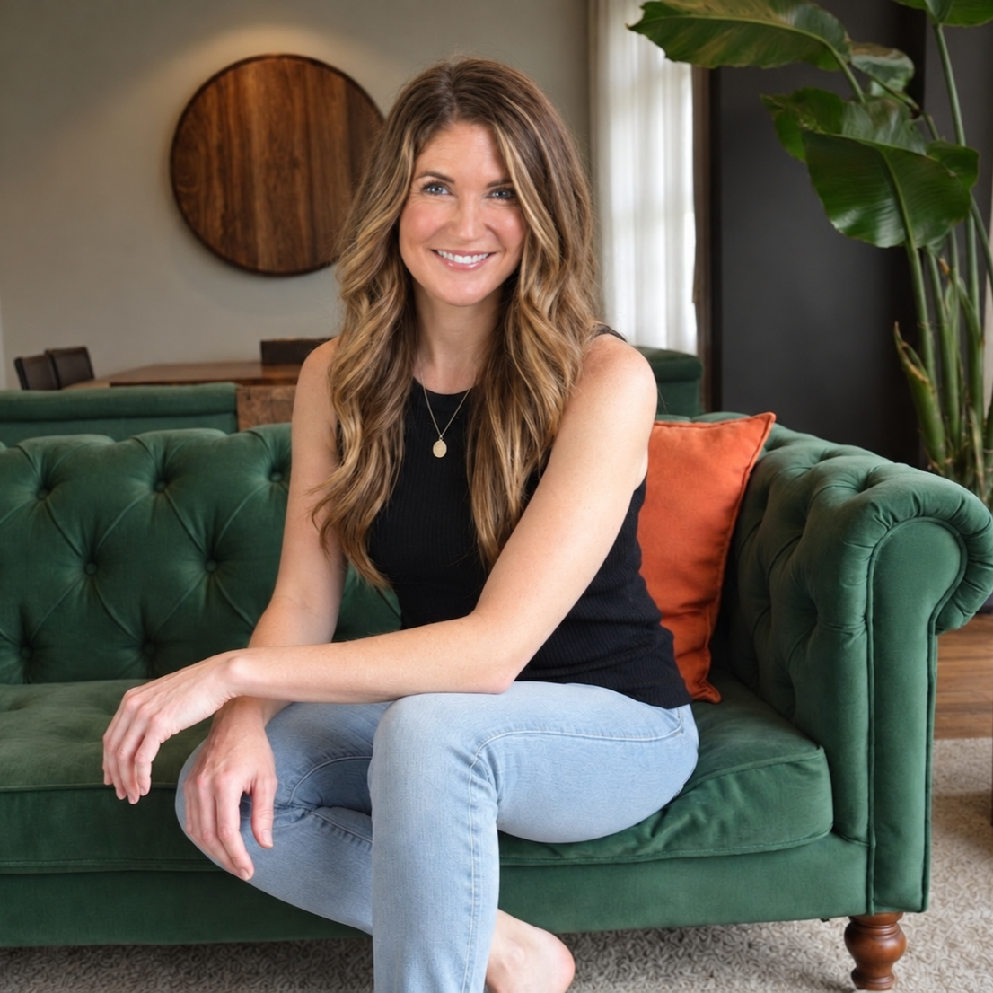 A woman with long wavy hair, wearing a black sleeveless top and light blue jeans, sitting on a green tufted sofa with an orange pillow in a cozy, well-decorated living room.