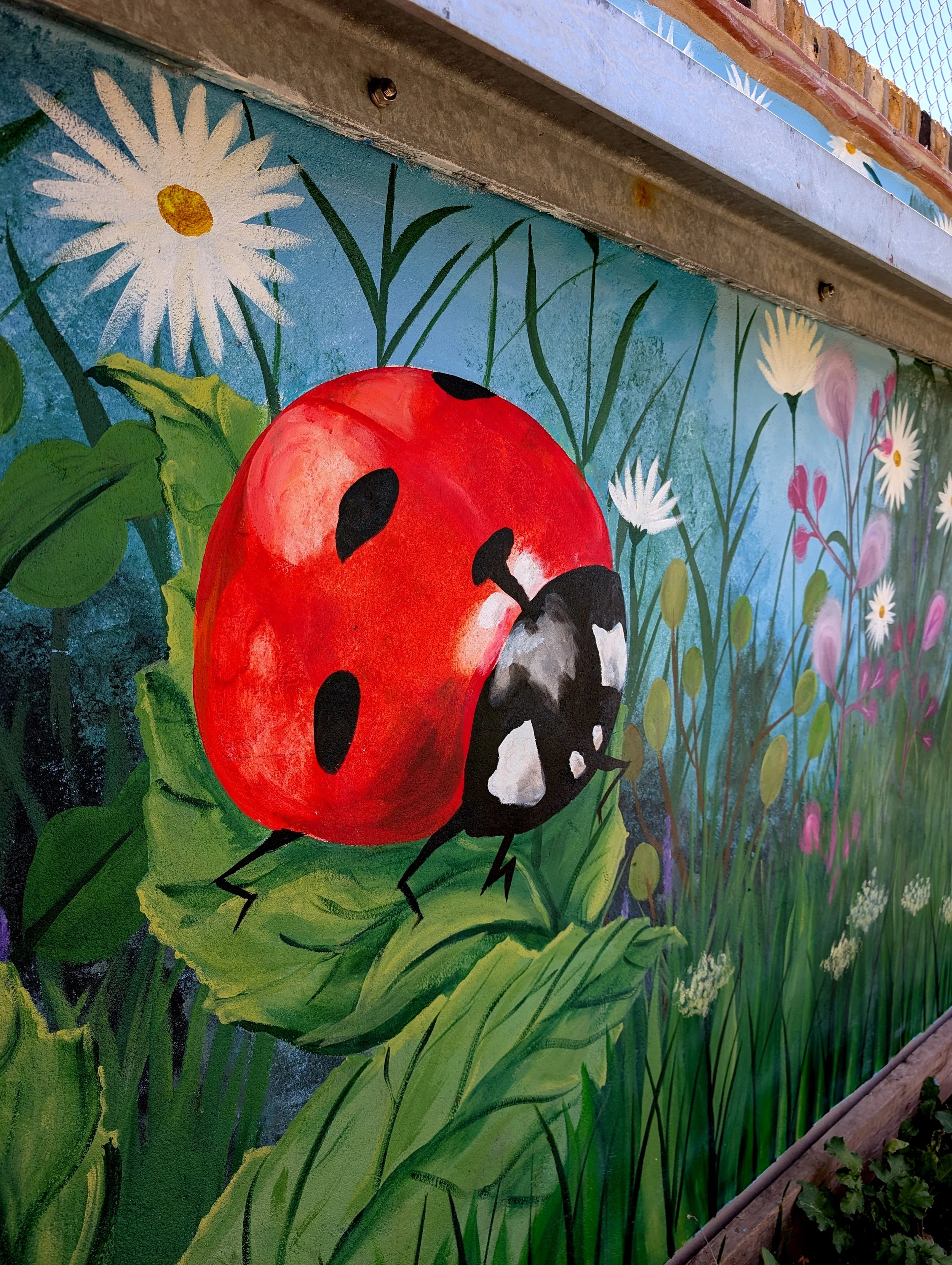 A colorful mural painting featuring a large red ladybug with black spots and black legs, surrounded by green grass, white daisies, pink flowers, and a blue sky background.