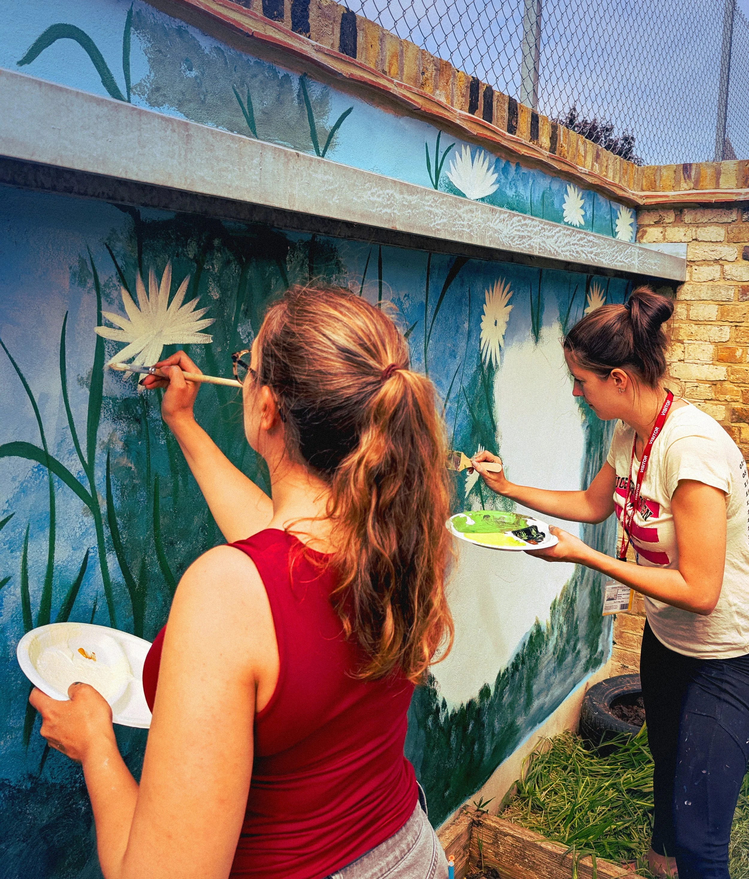 Two women painting a mural of white flowers with green stems on an outdoor wall. The woman in the foreground has red hair tied back and is holding a paintbrush and palette. The woman in the background has dark hair in a bun and is also painting.
