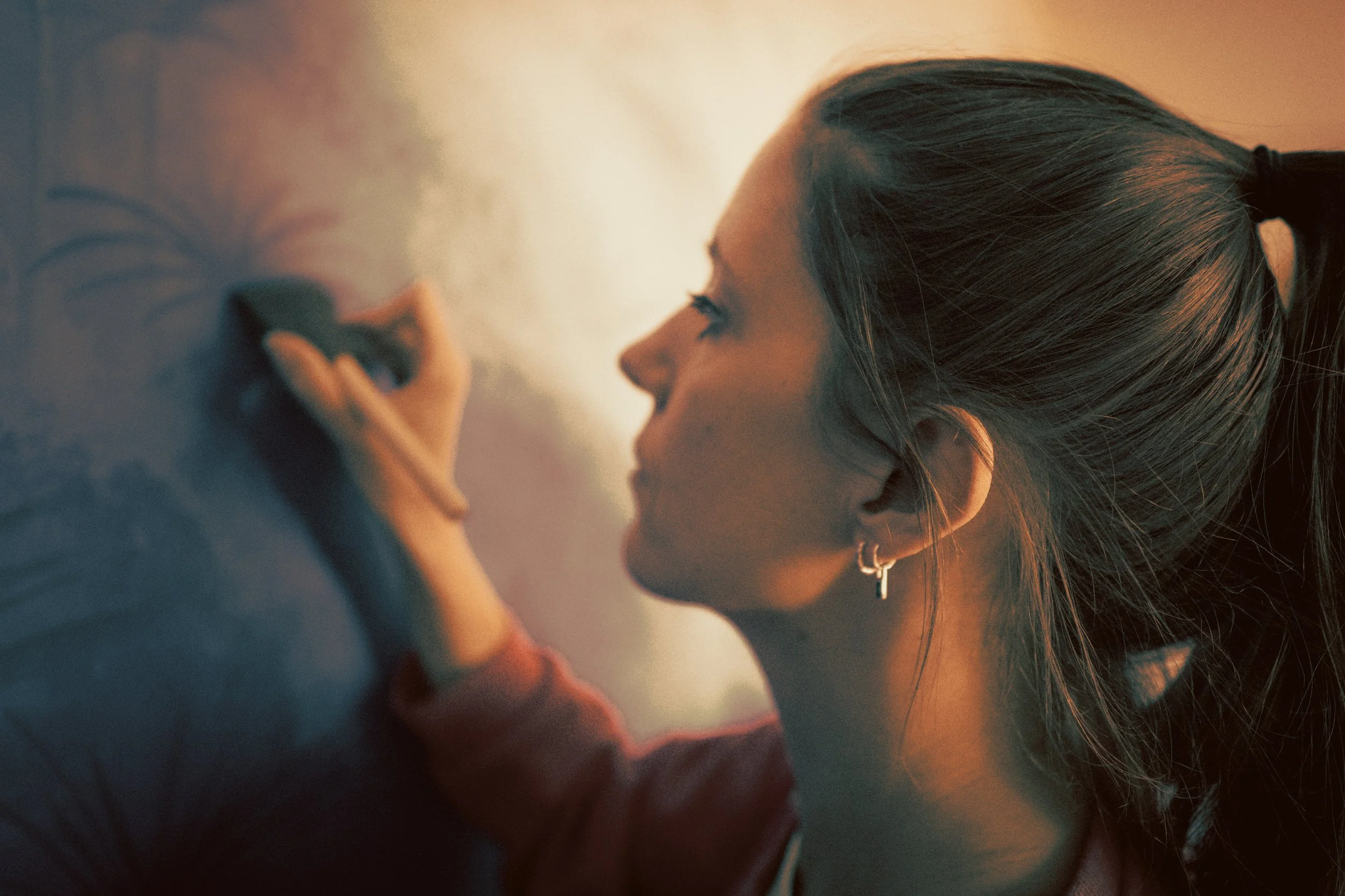 Young woman with long hair tied in a ponytail holding a makeup brush close to her face in a softly lit environment.