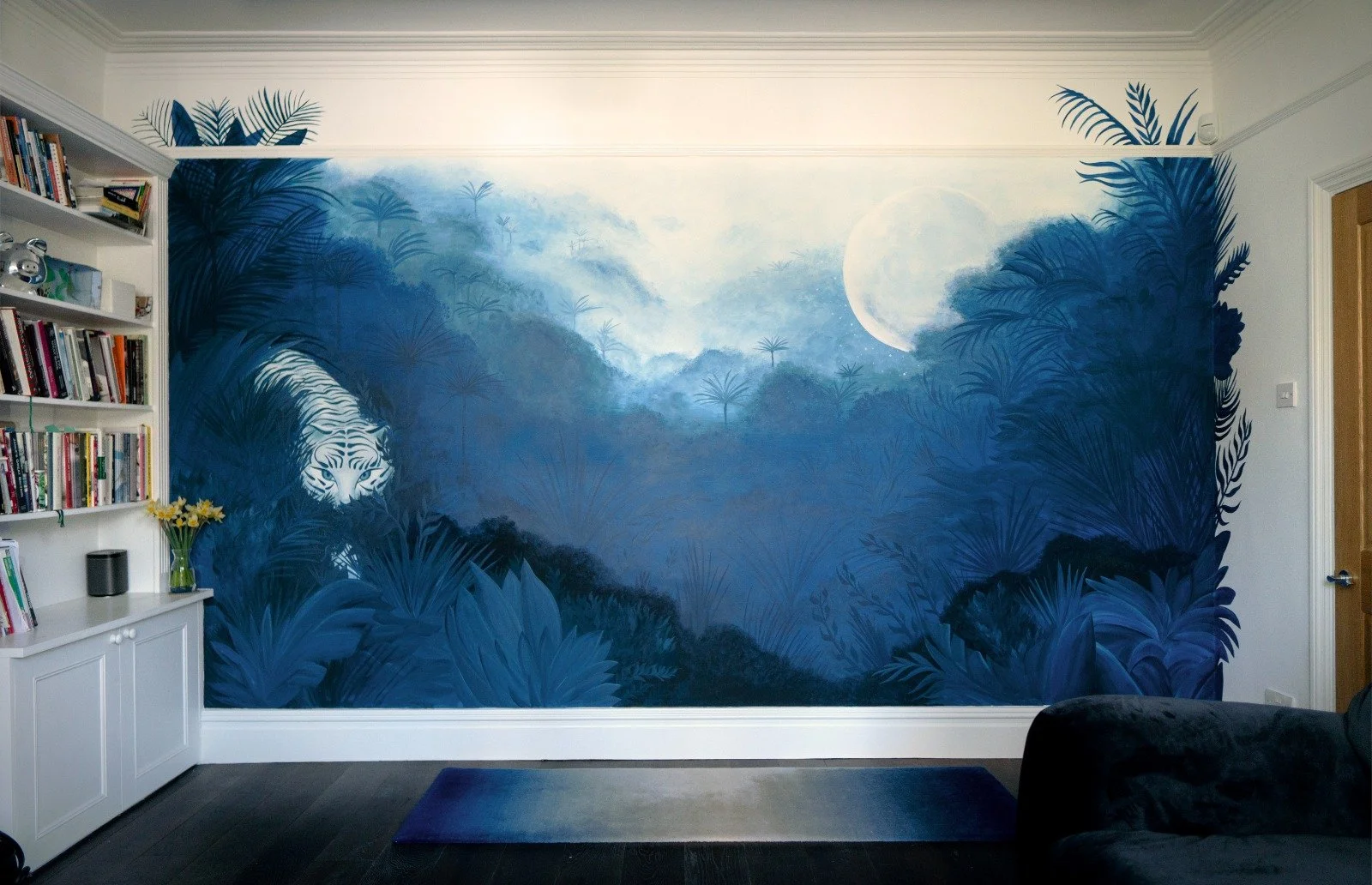 A living room with a large blue jungle mural painting of a white tiger hidden among lush foliage, with a white moon in the background.