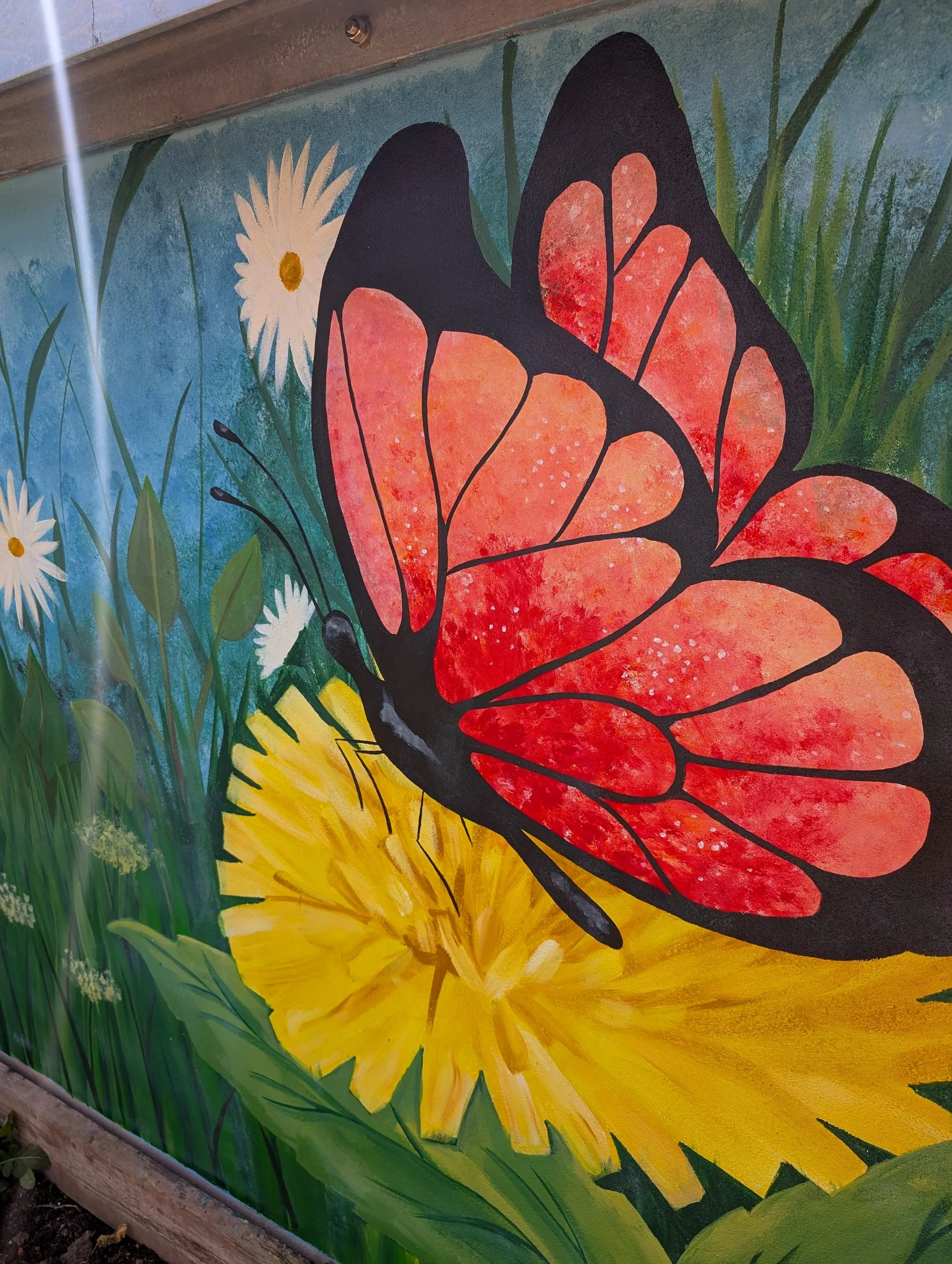 A colorful outdoor mural featuring a butterfly with orange and black wings, white daisies with yellow centers, green grass, and a bright yellow sun in a blue sky background.