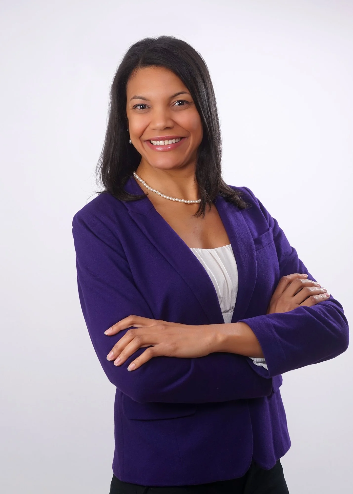 A professional woman with shoulder-length black hair, wearing a purple blazer, white blouse, pearl necklace, and earrings, smiling and crossing her arms against a plain white background.