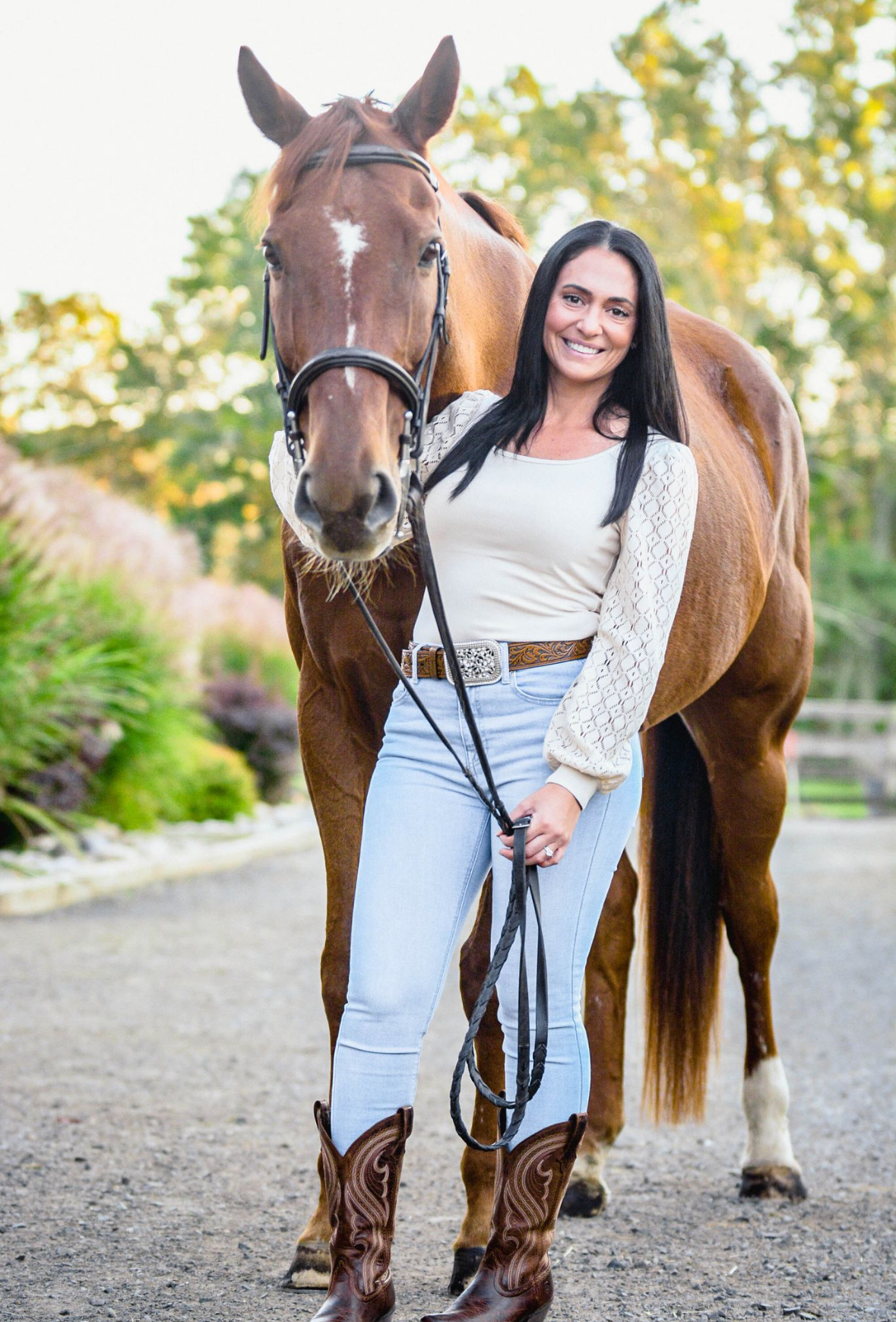 A woman standing with a brown horse outdoors, smiling, with trees and greenery in the background.